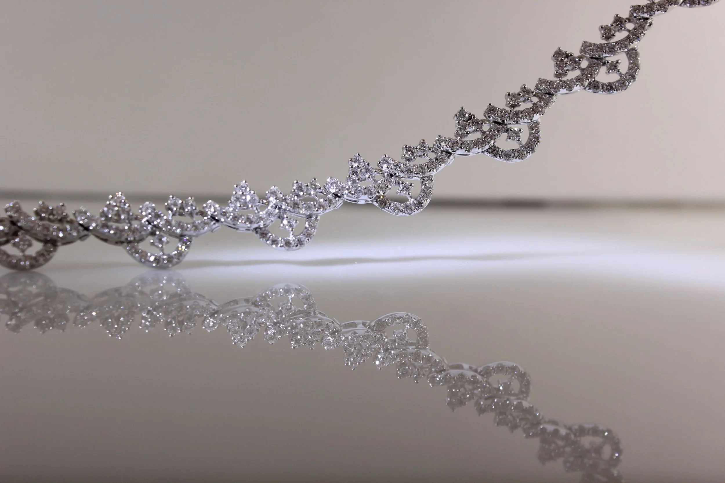 A close-up of a silver and diamond necklace on a reflective surface, with a soft light background highlighting the intricate details and sparkle of the jewelry.
