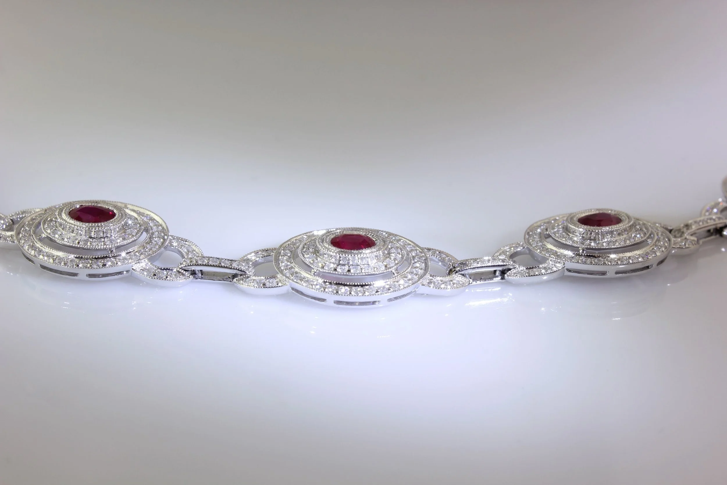 A silver bracelet with red gemstones and numerous small diamonds, resting on a white surface.