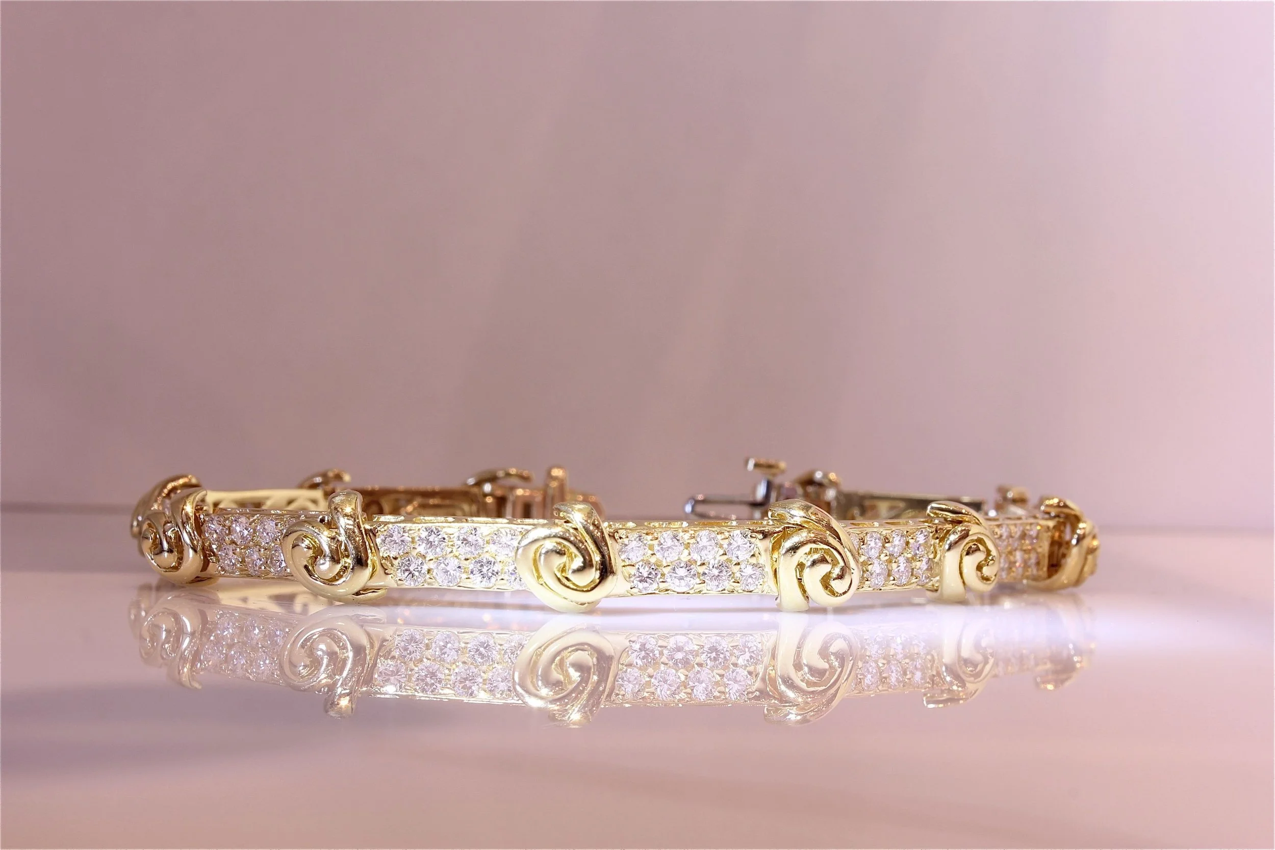 Gold bracelet with intricate scroll work and embedded diamonds, reflected on a shiny surface against a pink background.