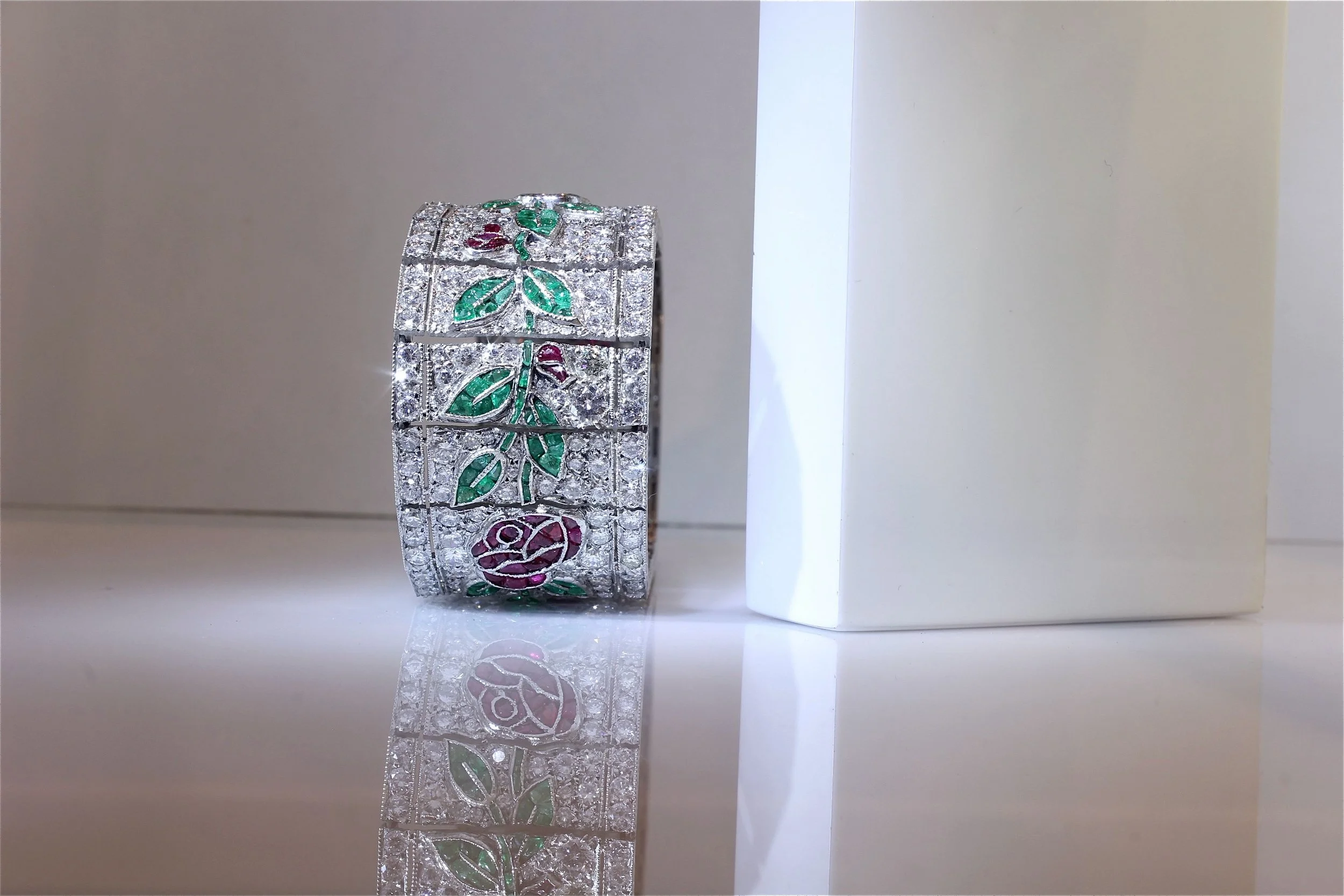 A silver bracelet with floral and leaf designs in red, green, and purple, embellished with crystals, reflecting on a shiny surface next to a white rectangular object.