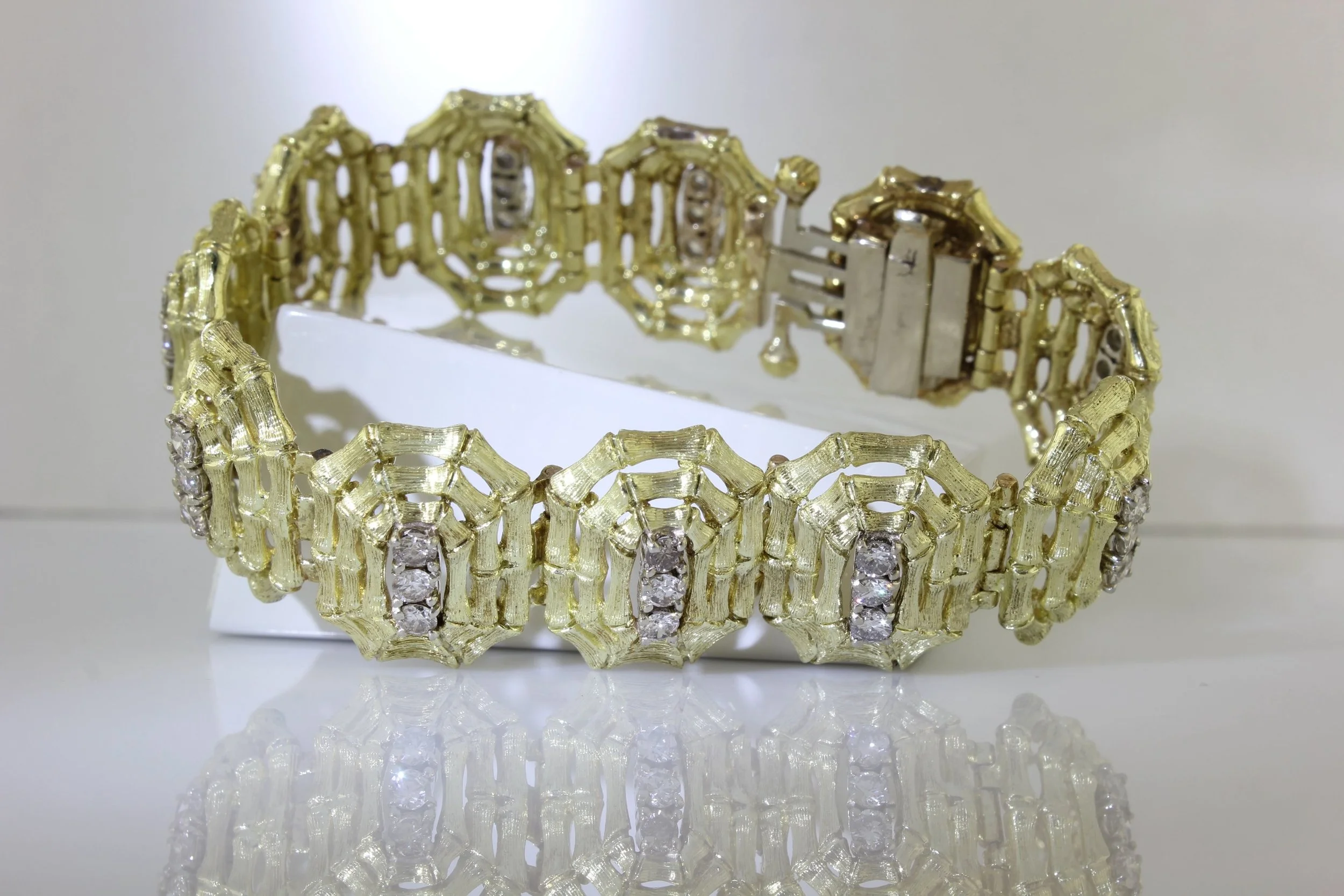 A gold bracelet with intricate design and embedded gemstones, displayed on a white stand with a reflection below.