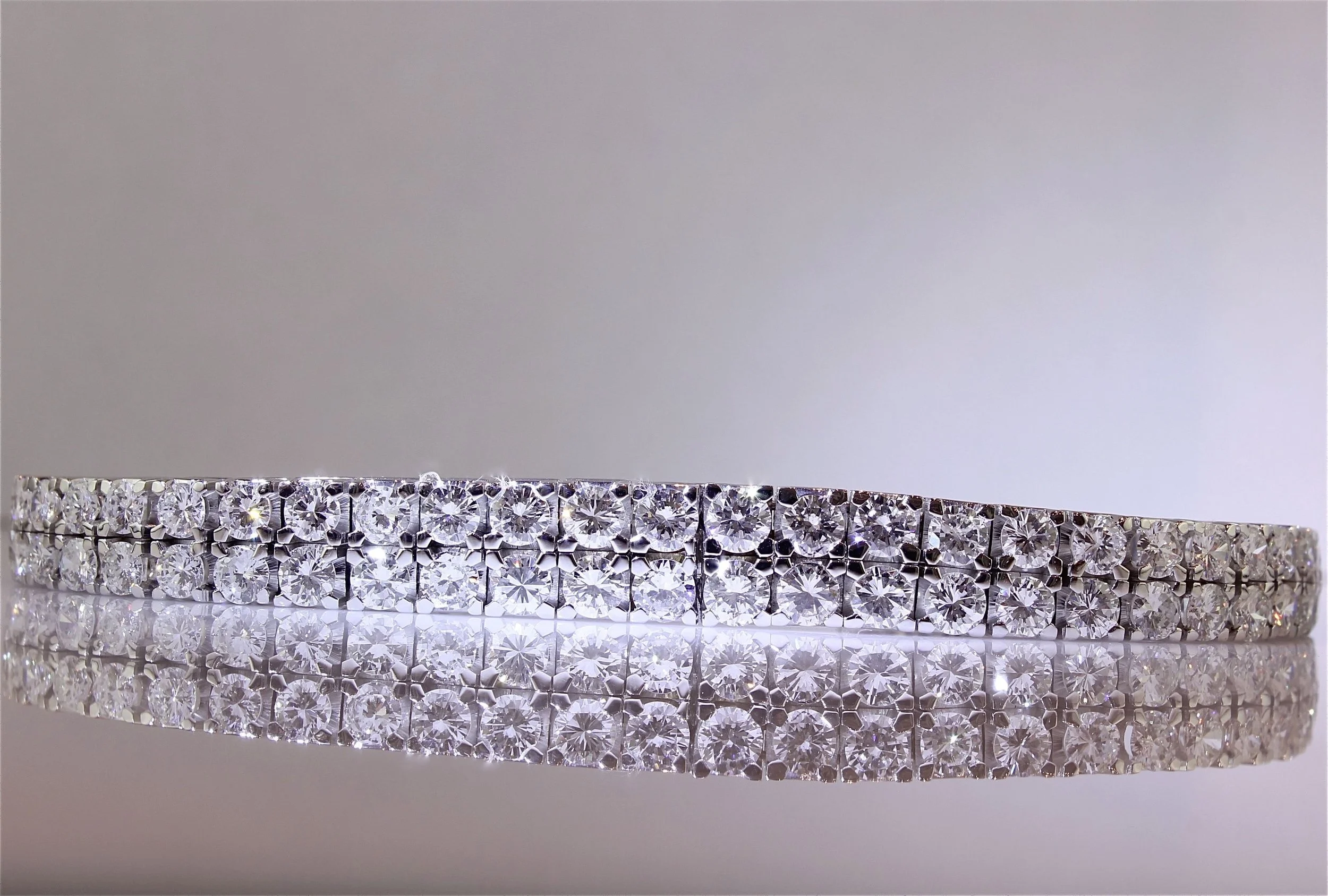Close-up of a diamond tennis bracelet with a precise row of sparkling, round-cut diamonds set in a metal band, reflecting light and placed against a white background.