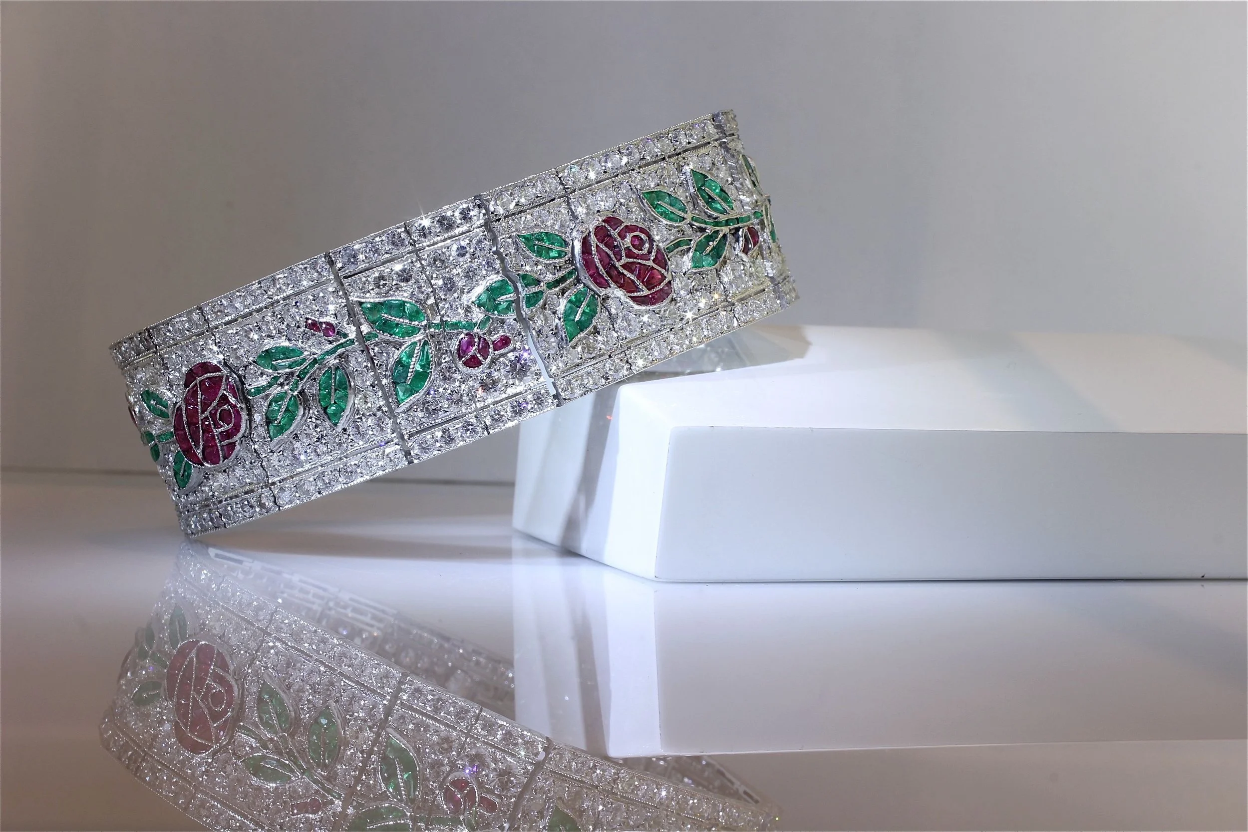 A decorative silver bracelet with embedded diamonds and painted floral design in red and green, displayed on a white stand with a reflective surface.