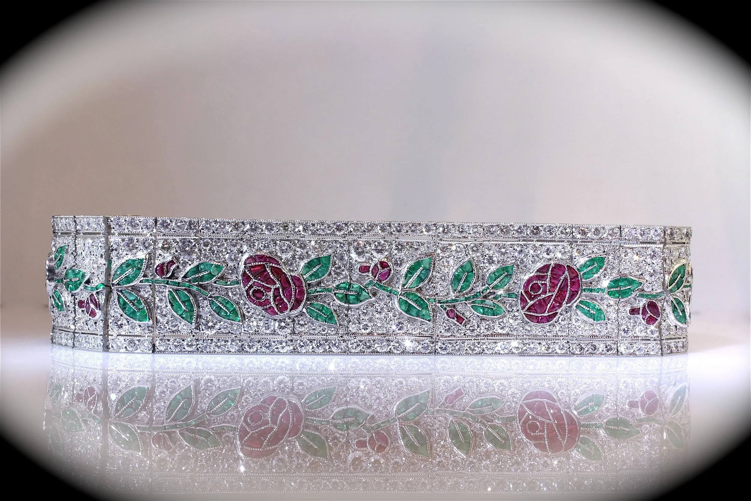 A close-up view of a silver bracelet with floral and leaf designs decorated with green and pink gemstones, reflected on a smooth surface.
