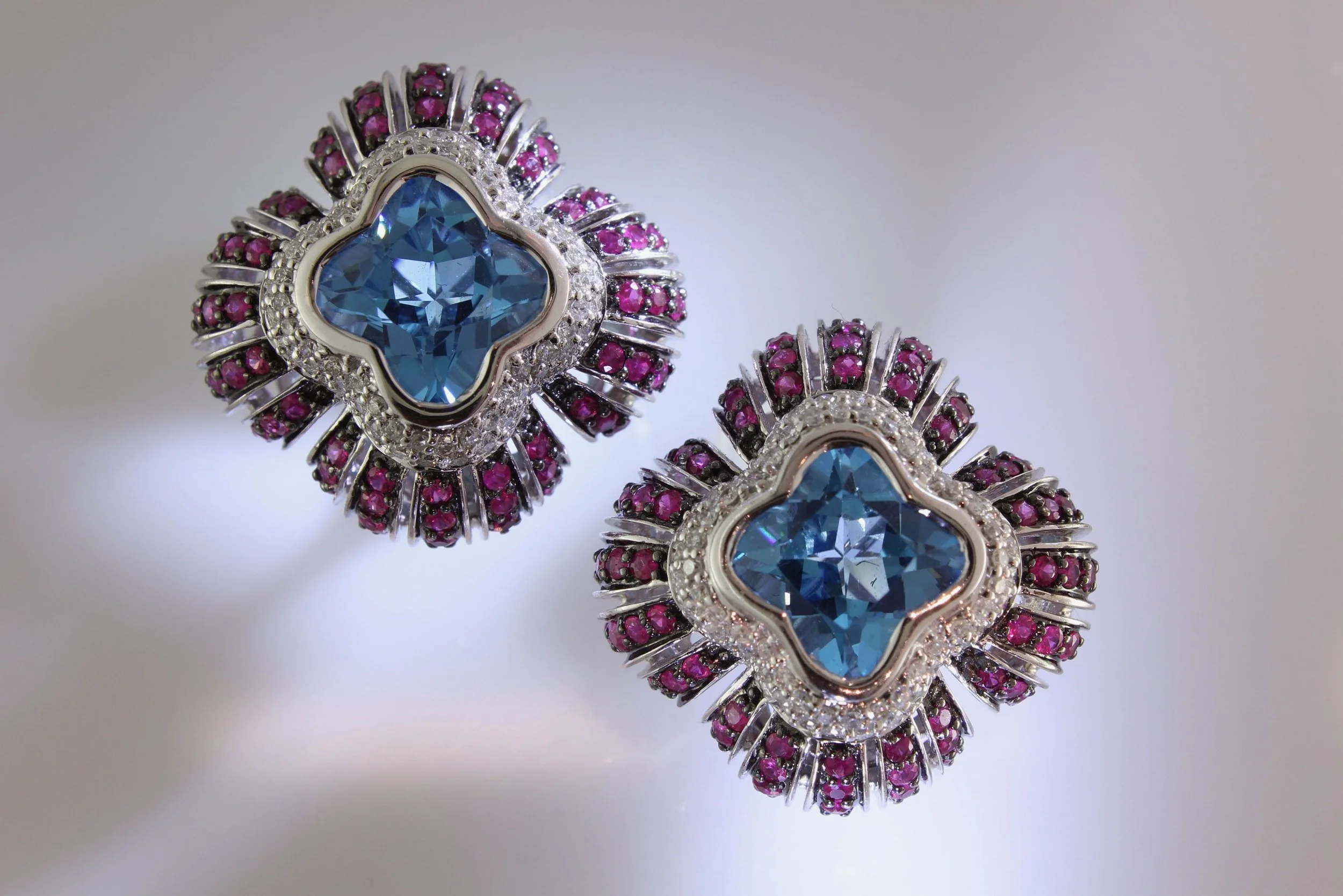 Jewelry earrings with large blue gemstones in the shape of a four-leaf clover, surrounded by small pink and clear diamonds.