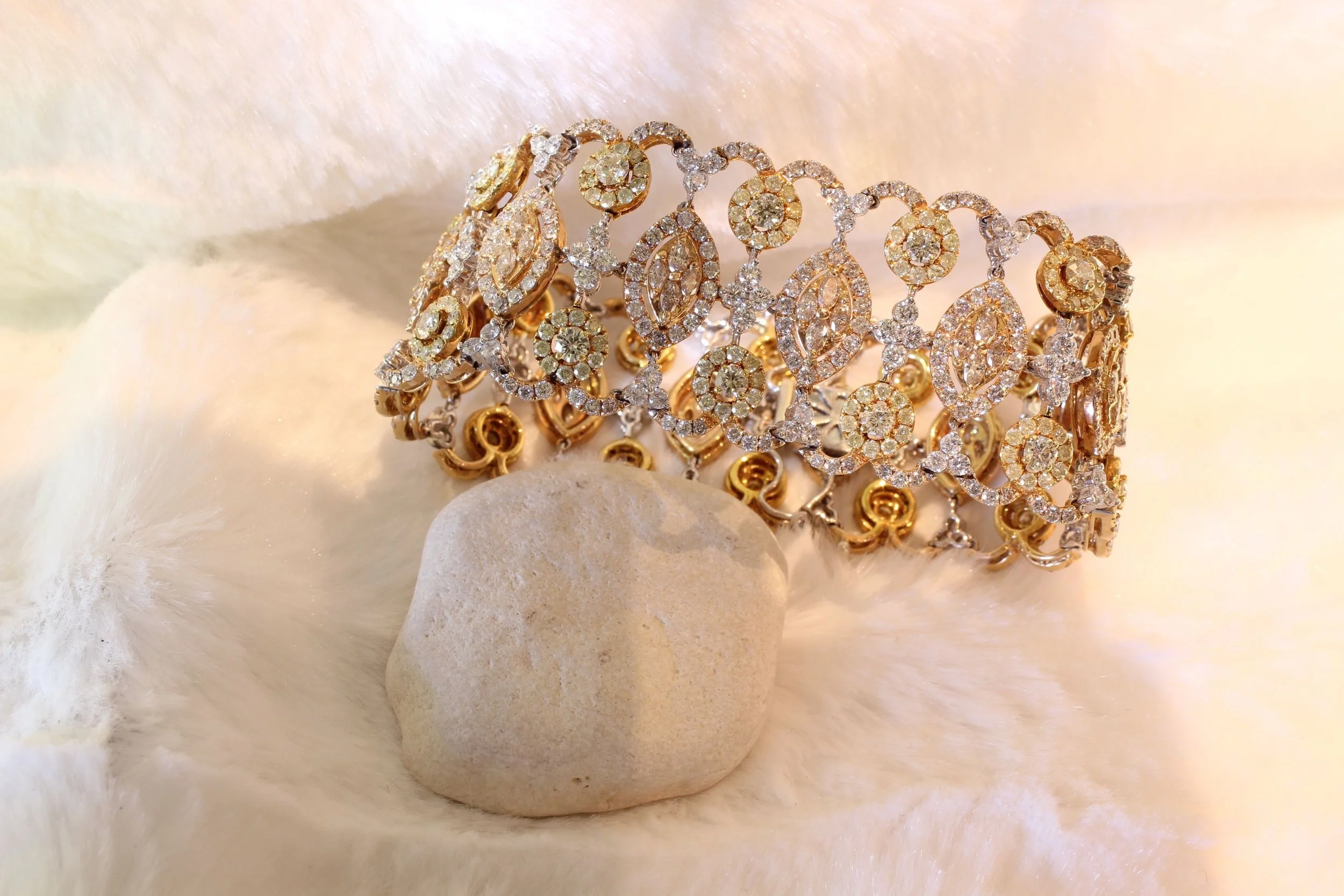 A gold and diamond jewelry bracelet with intricate design, resting on soft white fabric and light-colored stones.