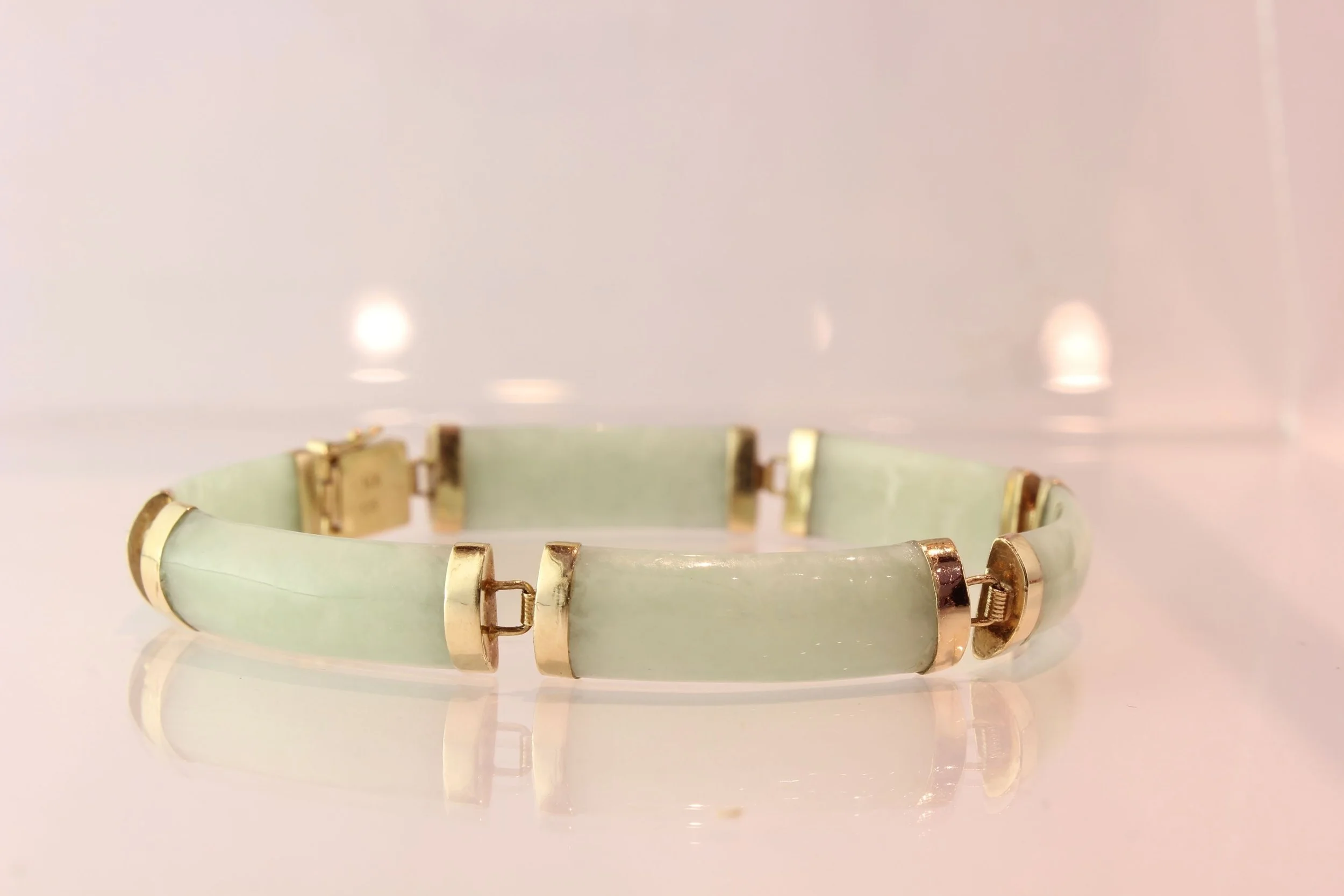 A jewelry bracelet with a pale green band and gold-colored rectangular accents on a reflective surface.