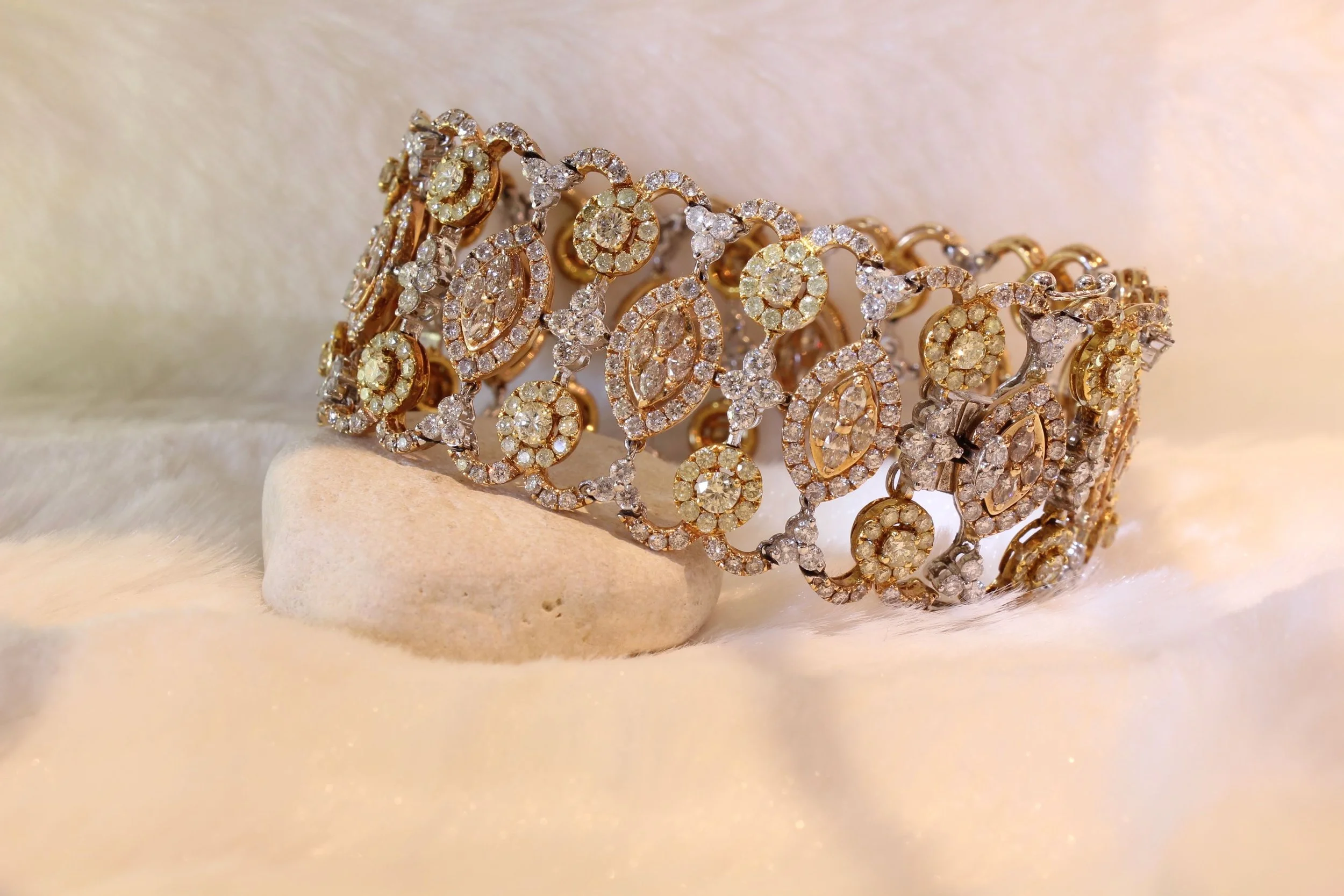 Gold and diamond jewelry bracelet resting on a beige stone with a soft fabric background.