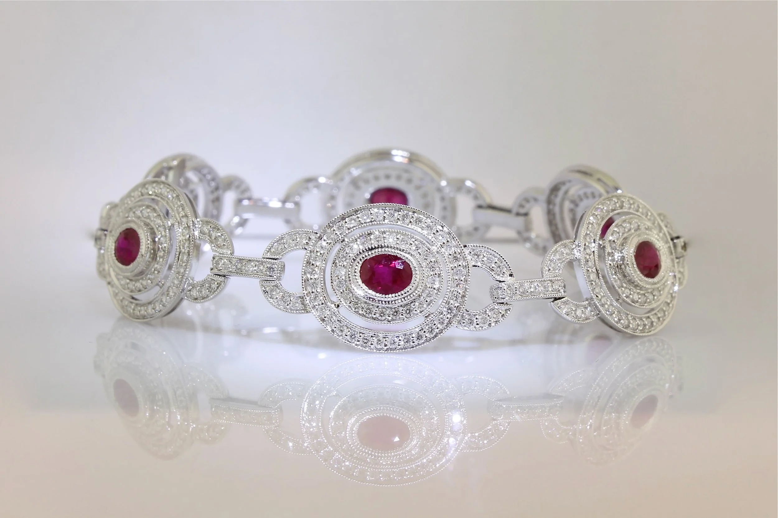 A silver bracelet decorated with multiple round-cut diamonds and a central large red ruby, featuring an intricate circular and geometric design, reflected on a glossy surface.