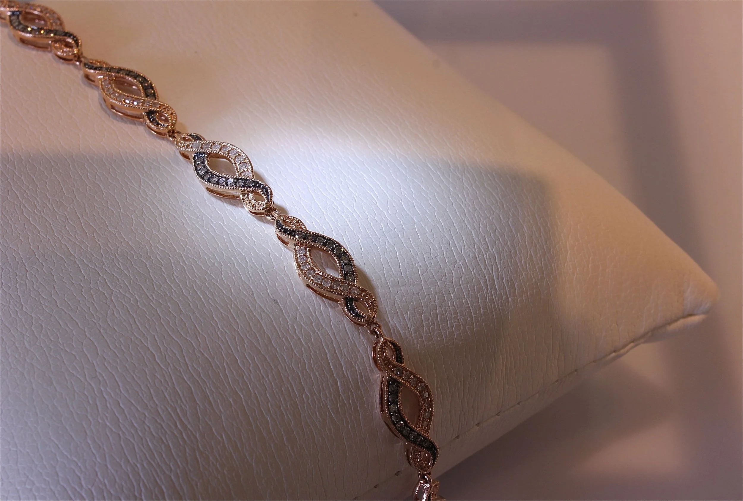 A gold-colored chain bracelet with intertwined loops decorated with small diamonds or crystals, displayed on a textured, light-colored jewelry stand.