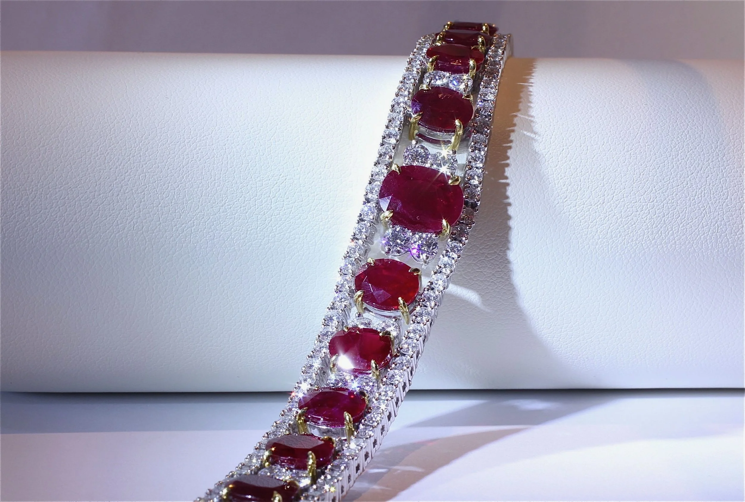 A silver bracelet with large red gemstones and numerous small clear diamonds, displayed on a white cylindrical surface.
