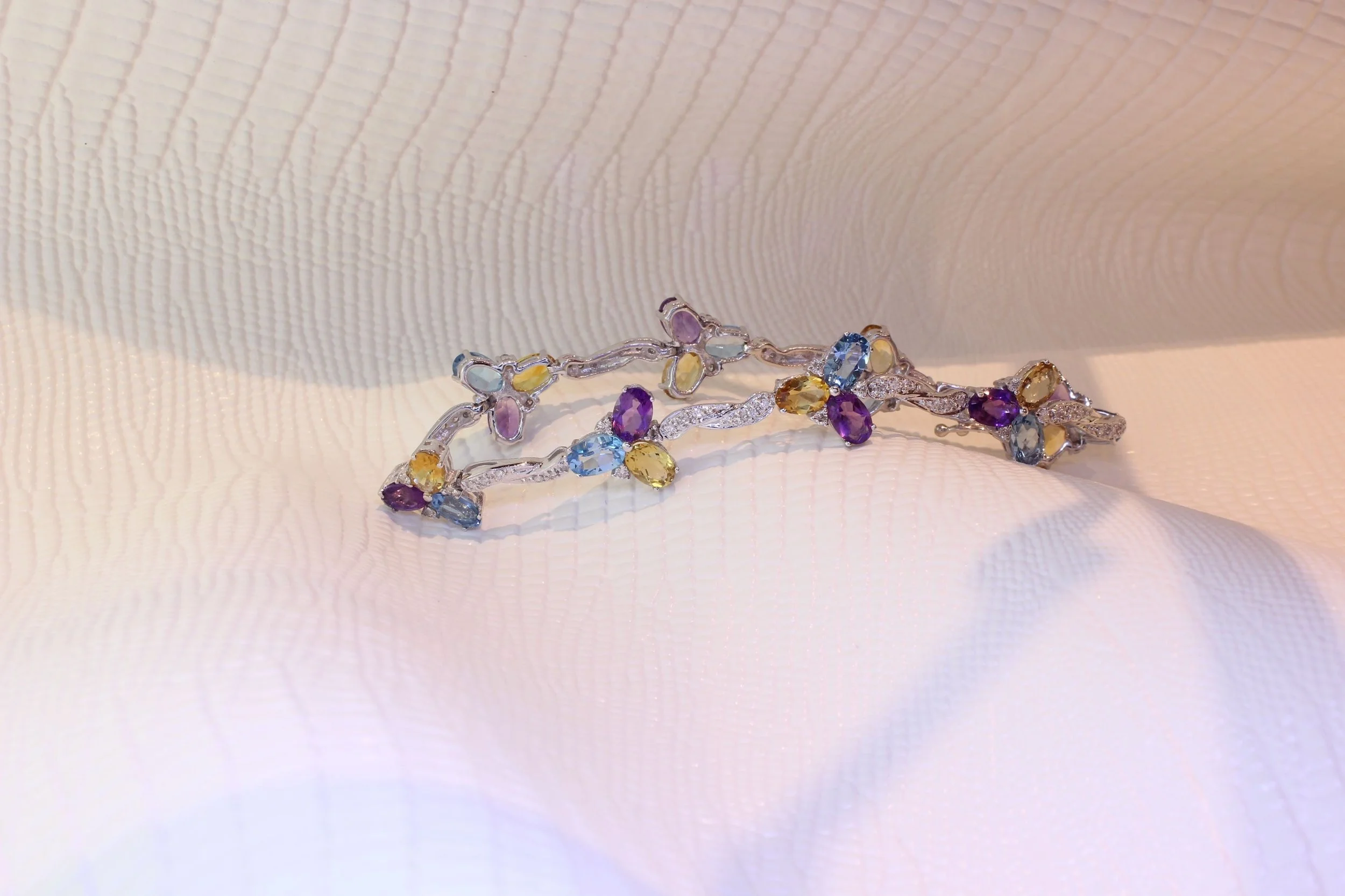 Two intricate silver bracelets adorned with multicolored gemstones, resting on a textured, cream-colored fabric.