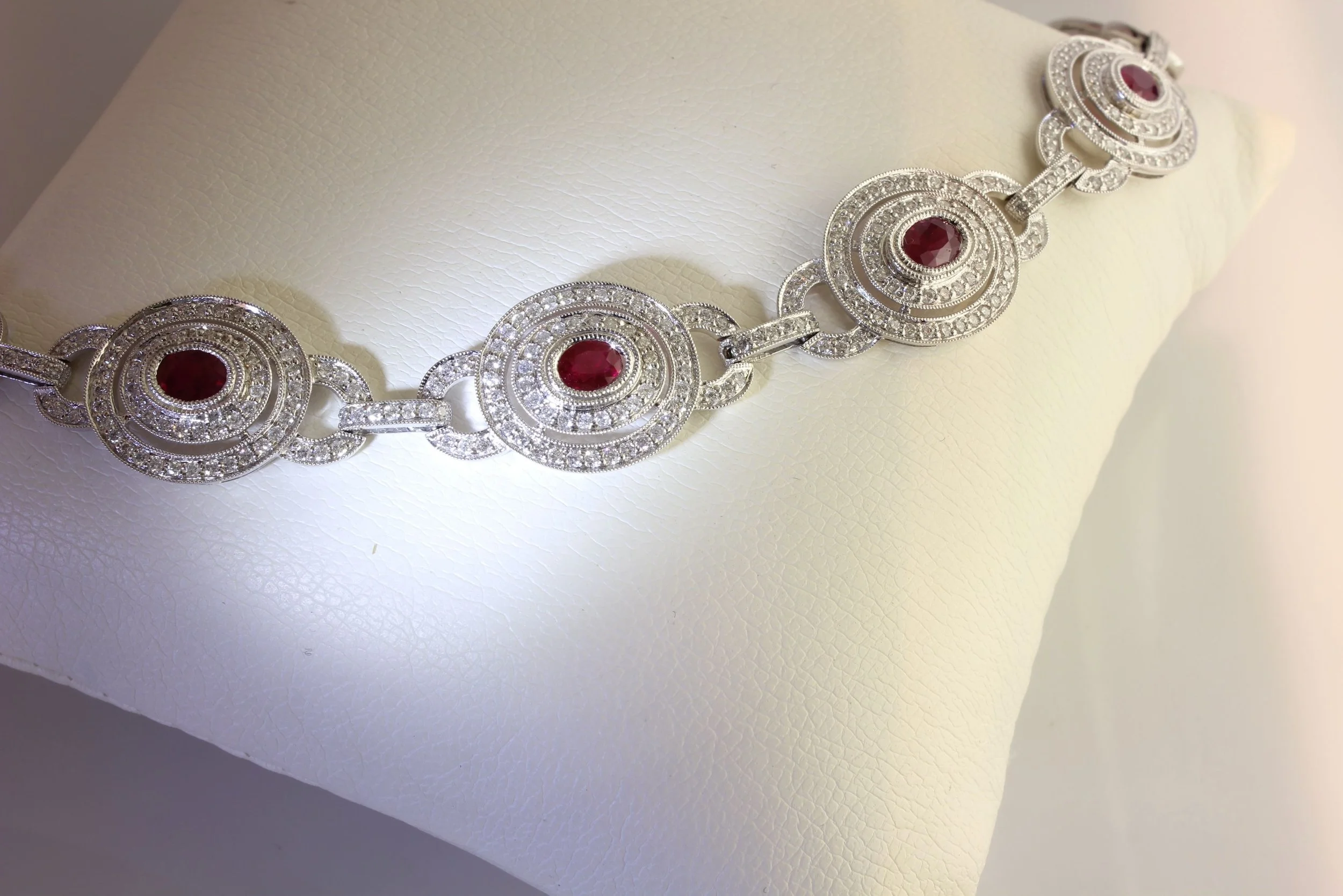 A silver necklace with circular patterns and red gemstones, displayed on a white textured jewelry holder.