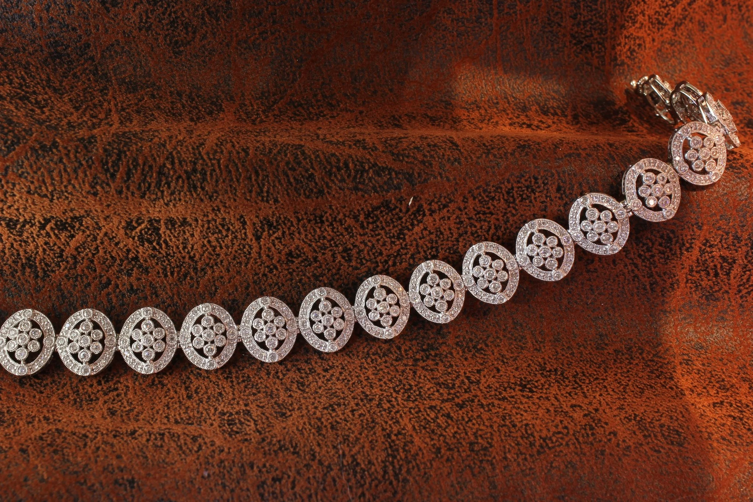 A white gold bracelet with circular and oval-shaped designs encrusted with small diamonds, resting on a textured reddish-brown velvet fabric.