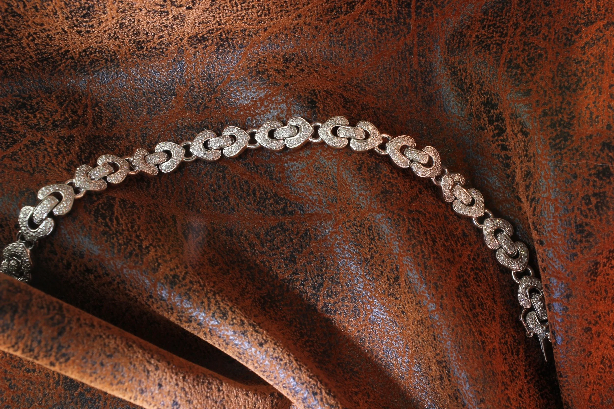 A white gold bracelet with interconnected links, each decorated with small sparkling stones, resting on a textured, rust-colored fabric.