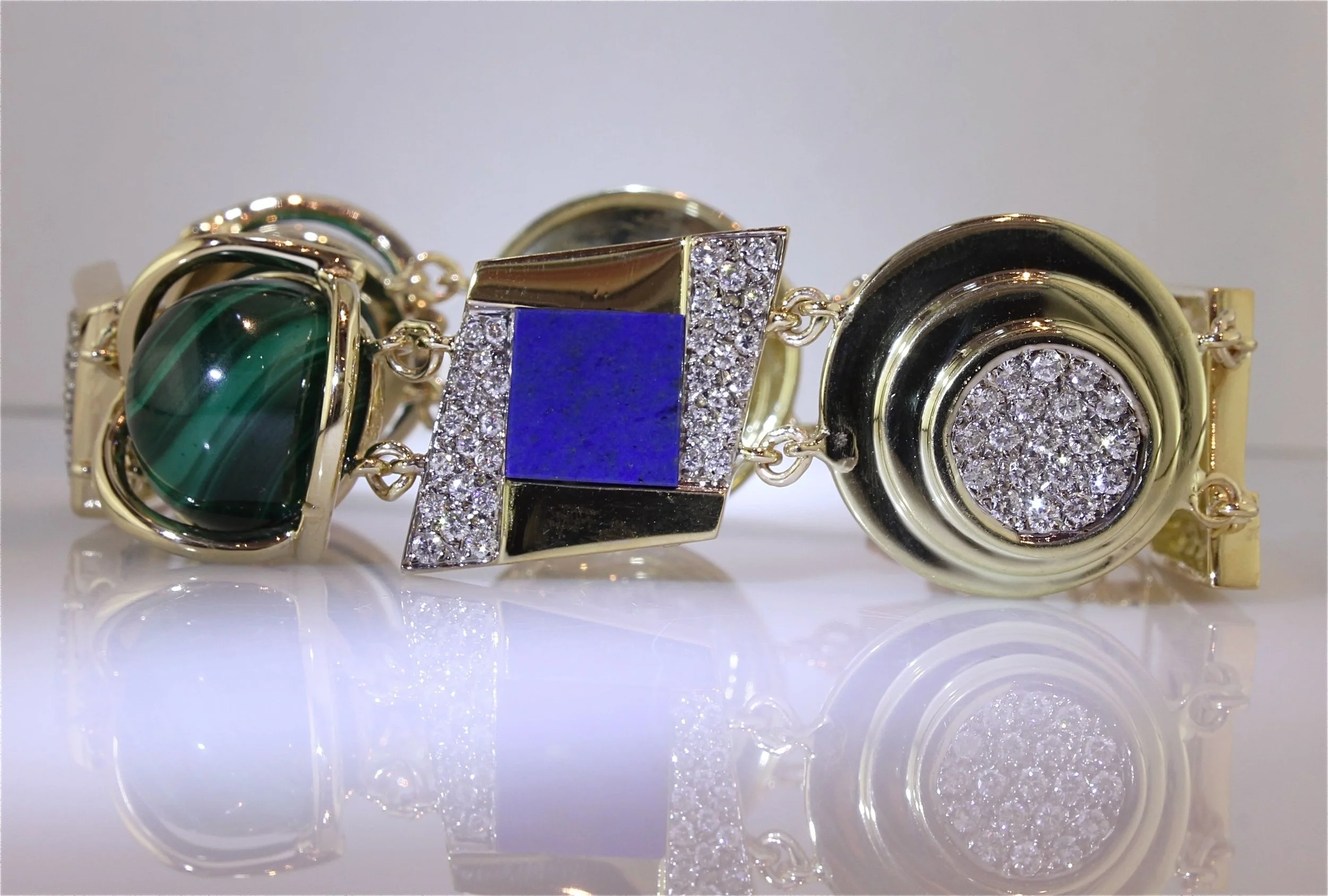 Gold bracelet with large oval green stone and square blue stone, decorated with numerous small diamonds, on a reflective white surface.