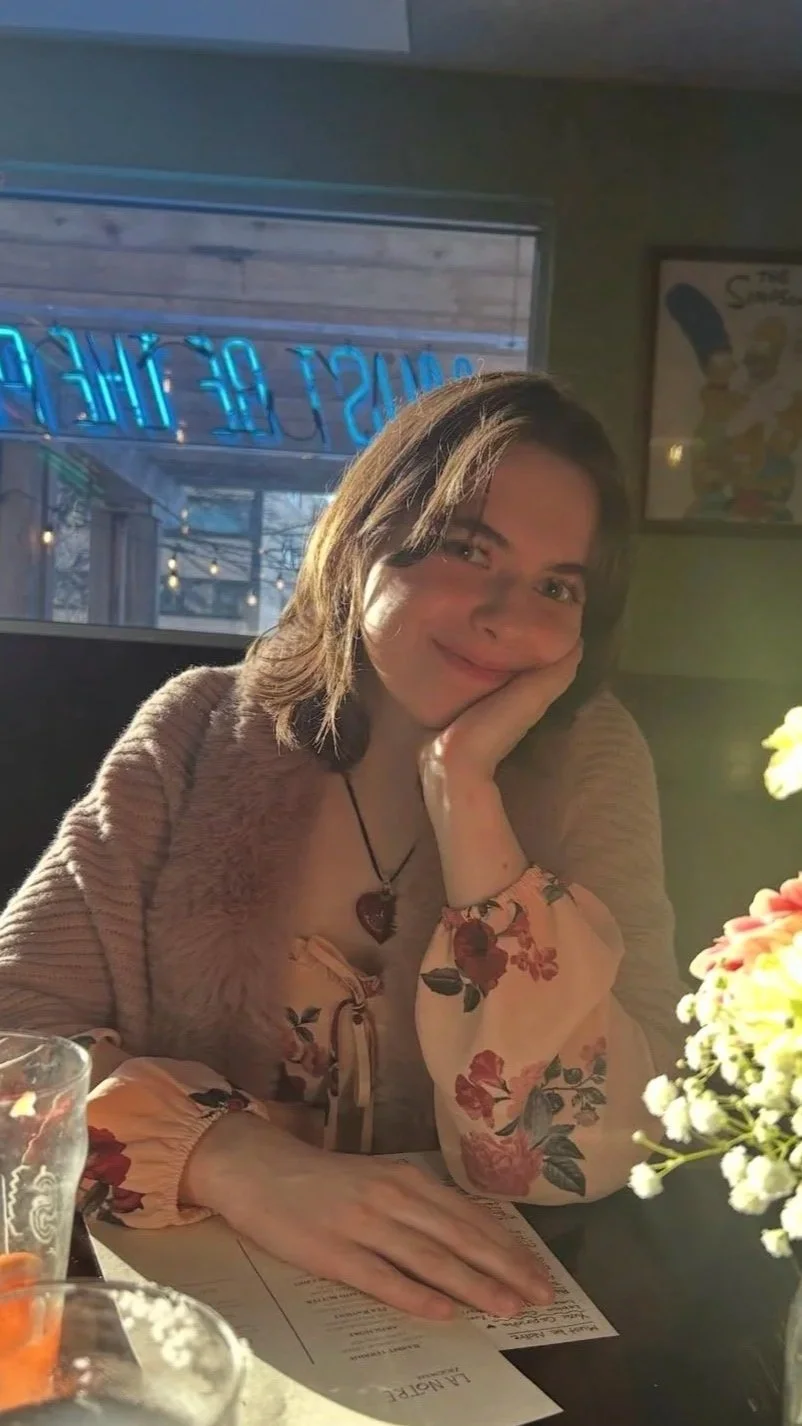 Ava sits at a table inside a restaurant. She has shoulder length brown hair and is wearing a brown sweater over a floral blouse. Her face is resting in her hand as she smiles at the camera.