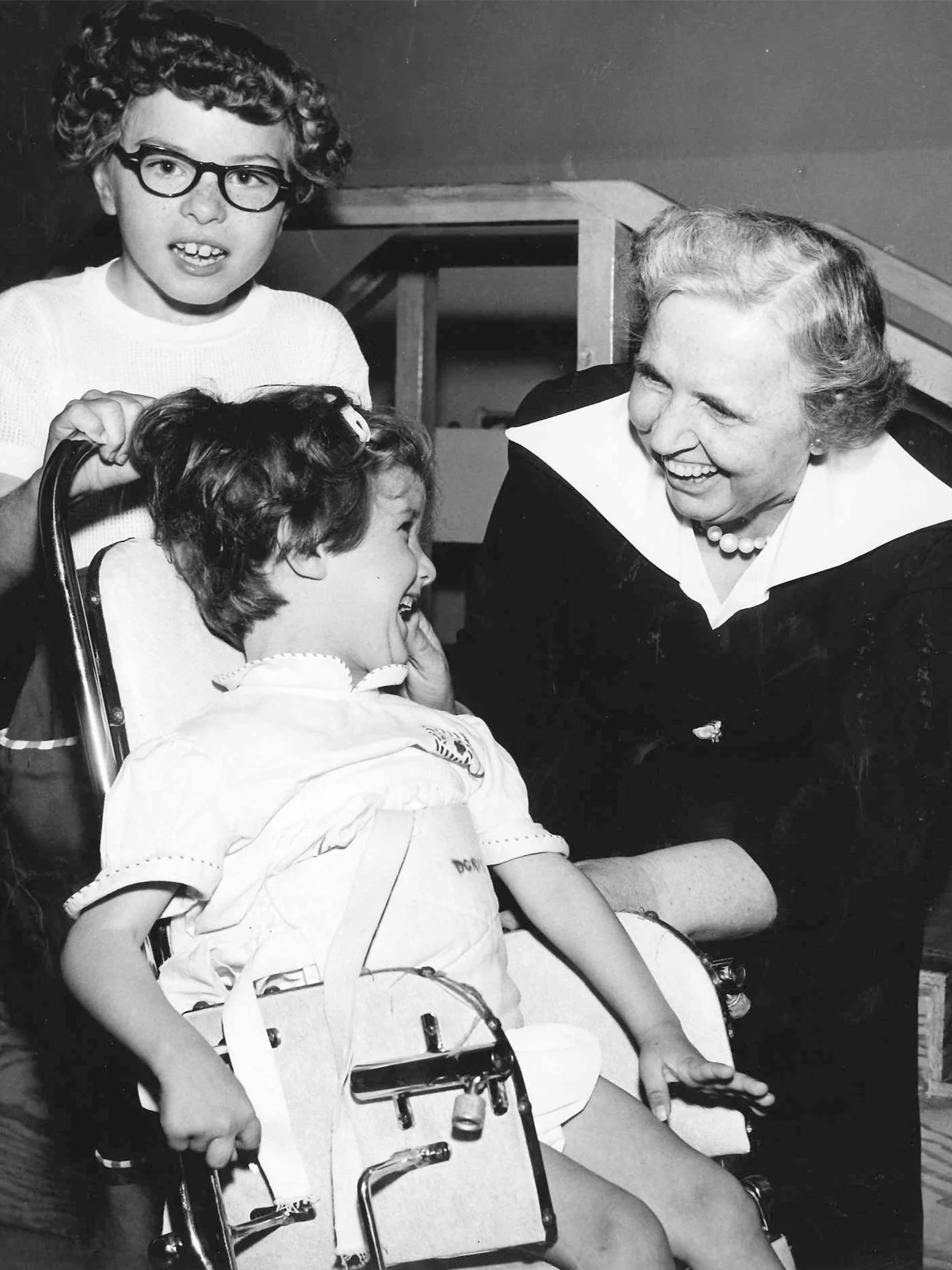 An old black and white photo. An elderly woman wearing pearls and a black dress is looking at a young girl in an old-fashioned wheelchair. The young girl is smiling back. Behind her another young girl stands with her hands on the back of the chair.