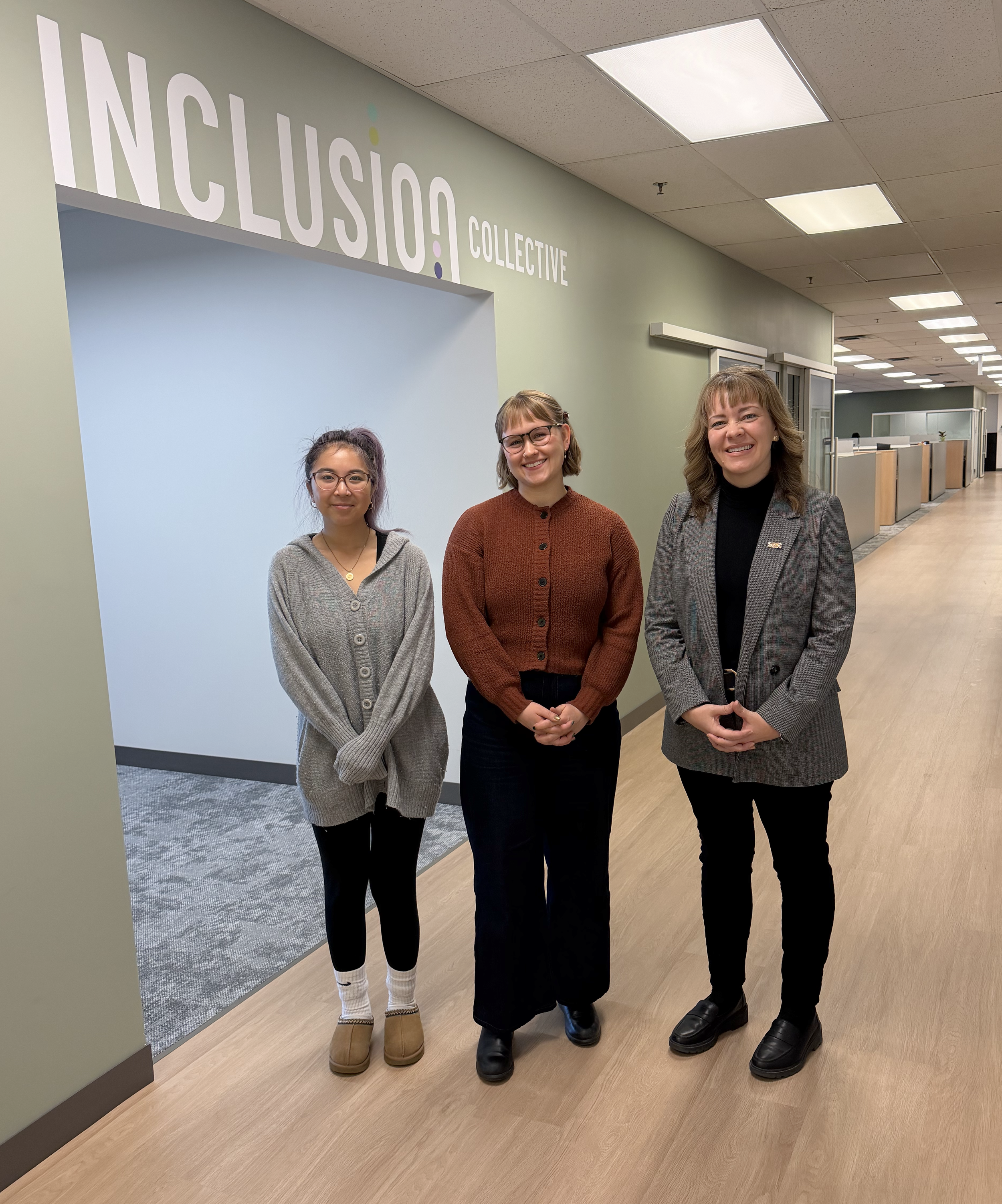 Three women stand posing for a photo inside Manitoba Possible. Behind them is a large sign that reads 'Inclusion Collective'