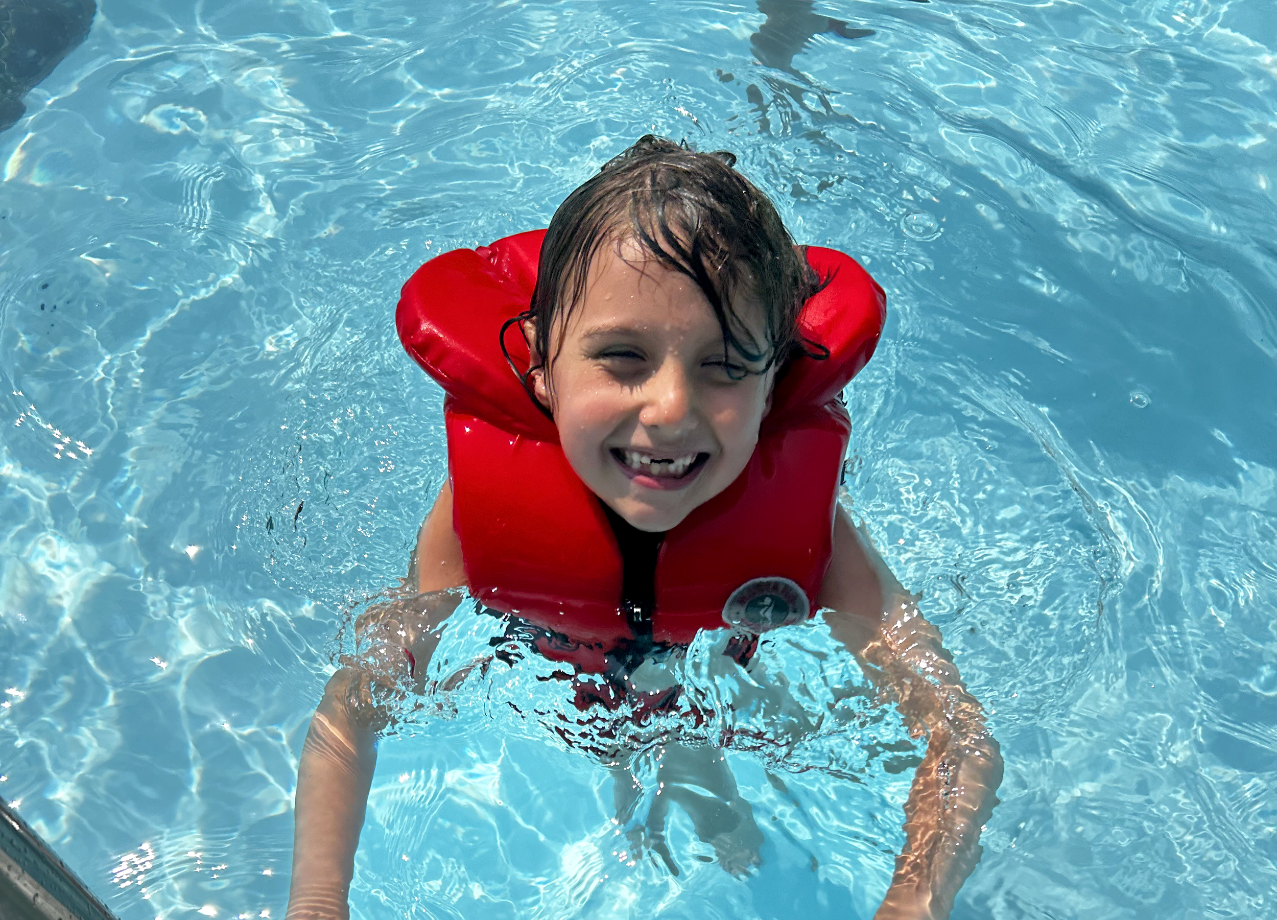 A young child is wearing a life jack in a pool. It appears to be a warm sunny day.