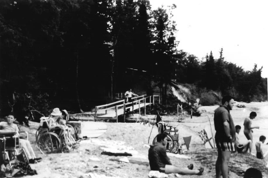 An old black and white photo of people lounging on a sandy beach by the water. some are wheelchair users and some are sitting and and standing.