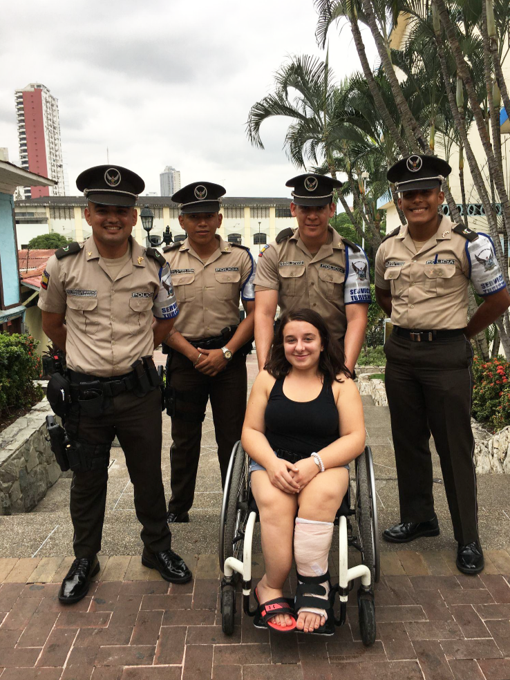 Meagan poses for a photo with the four police officers who carried her up steep steps in Ecuador. They are all dressed in police uniform. Meagan is sitting in her wheelchair.