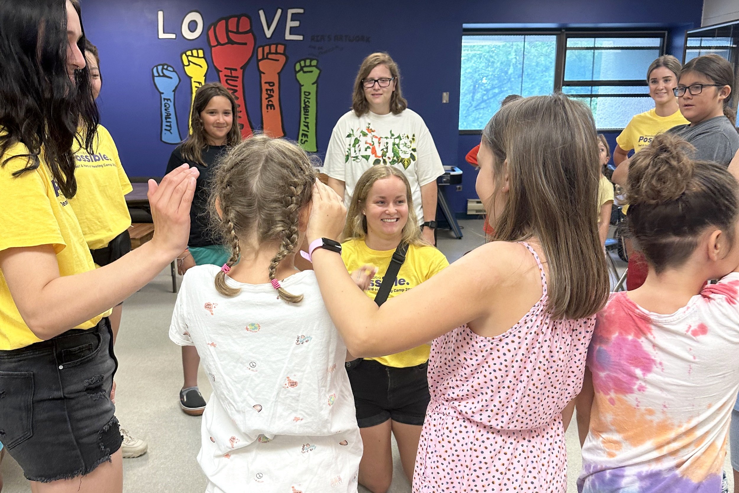 A camp counsellor with long blonde hair is smiling at two girls who are playing a game.