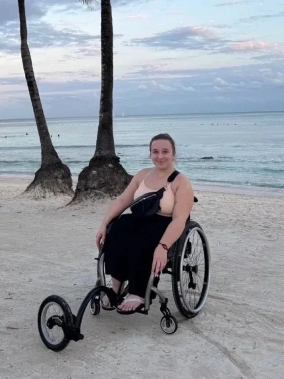 A photo of Meagan on a beach. Behind her is bright blue water and tall palm trees. Meagan has light brown hair in a pony tail. She is a wheelchair user, with a special third wheel attachment on the front of her wheelchair.