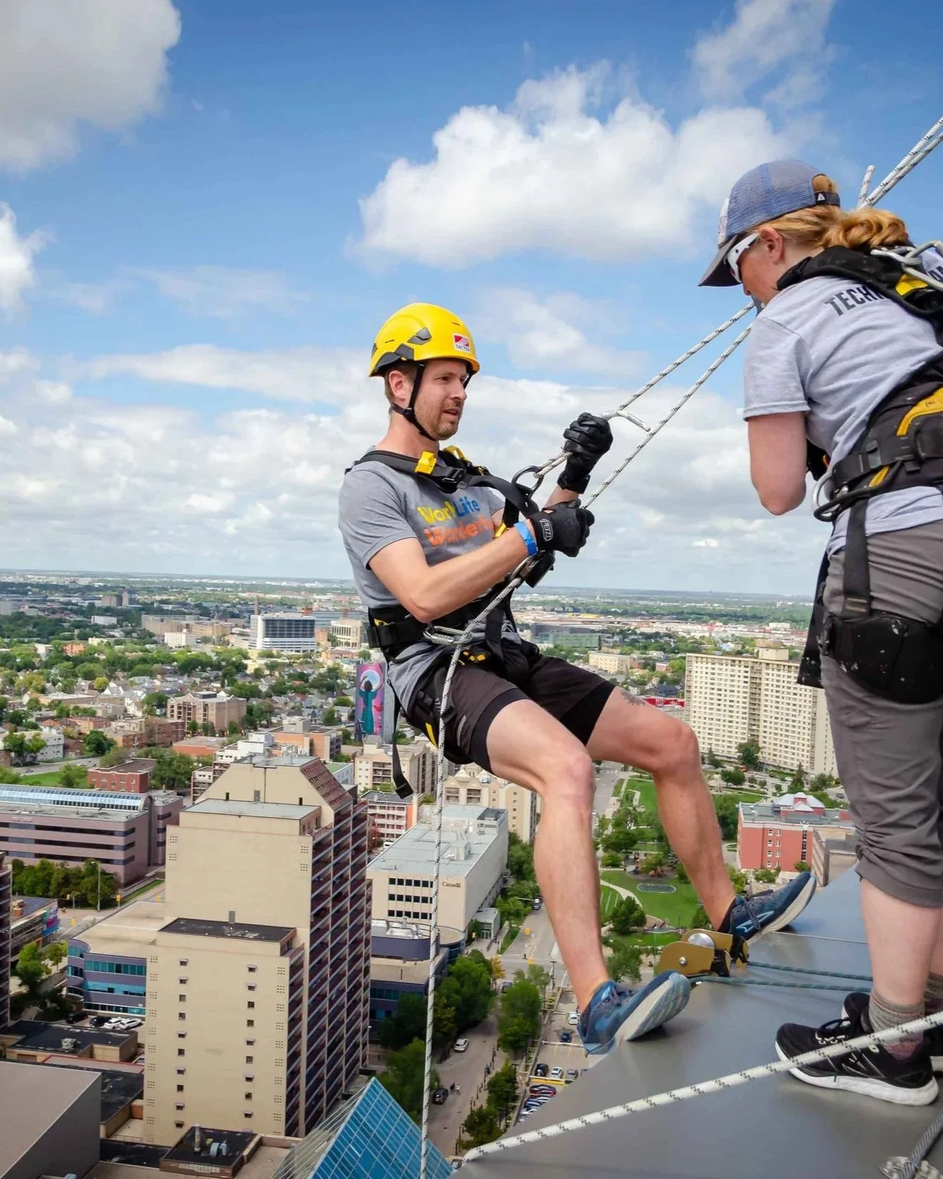 A man with harnesses, ropes and a hard hat leans off the side of the roof of Manitoba Hydro. The tops of apartment buildings are below him and a bright blue sky in the background.