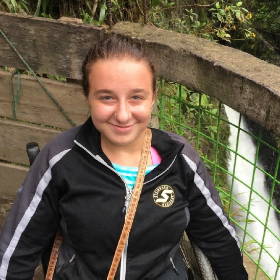 Meagan in front of a waterfall in Ecuador. She is wearing her hair in a ponytail and a black and gray jacket.