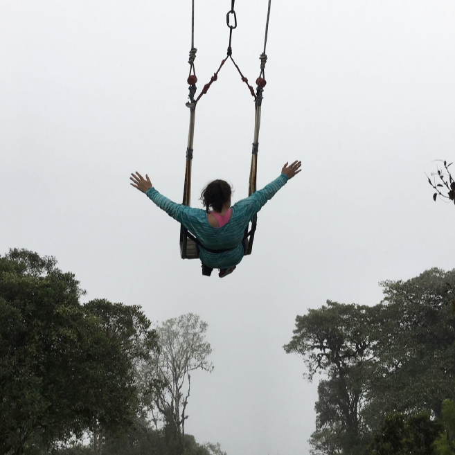 Meagan is harnessesd into in a swing with her arms reaching into the air. She appears to be very high in the air as all you can see below her  are tree tops.