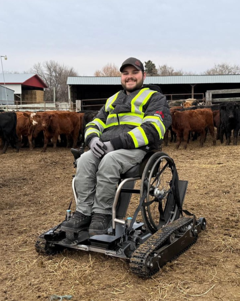 a young man who is a wheelchair user poses for a photo on a farm. He has a special all-terrain attachment on his manual wheelchair. He is wearing a reflective work jacket and hat. behind him are dozens of cows.