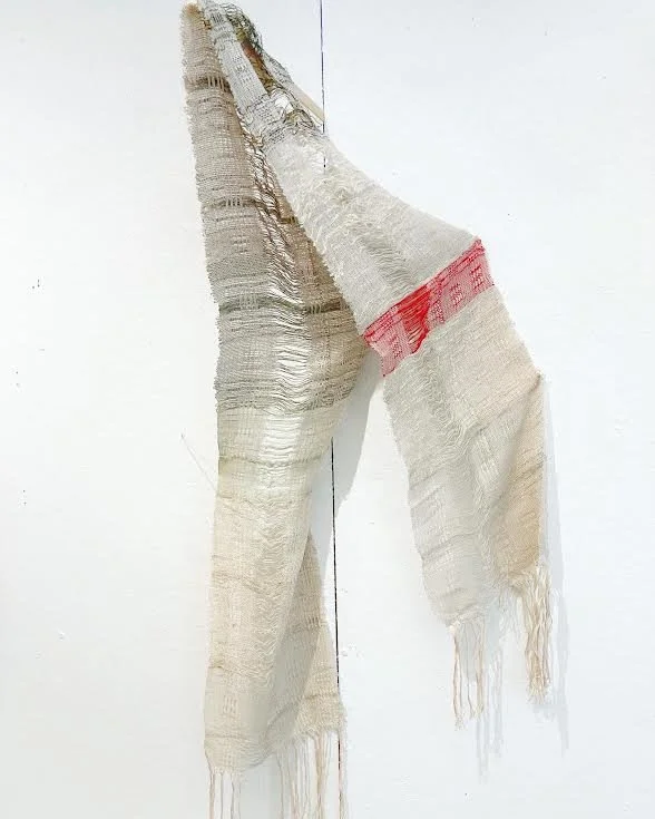 A wadded, beige fabric with frayed edges hanging on a white wall, with some red pattern or marking near the top.