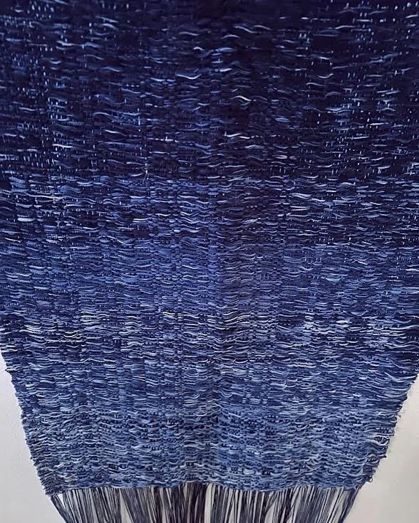 Close-up of a textured, knitted fabric or textile in shades of blue and purple with a fringed bottom edge.