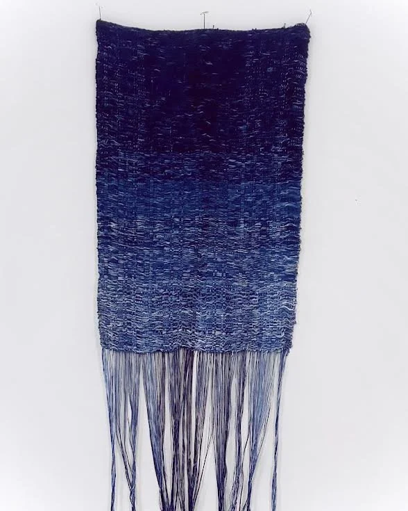 A woven textile artwork hanging on a wall with a gradient of dark blue to light blue, featuring long dangling threads at the bottom.