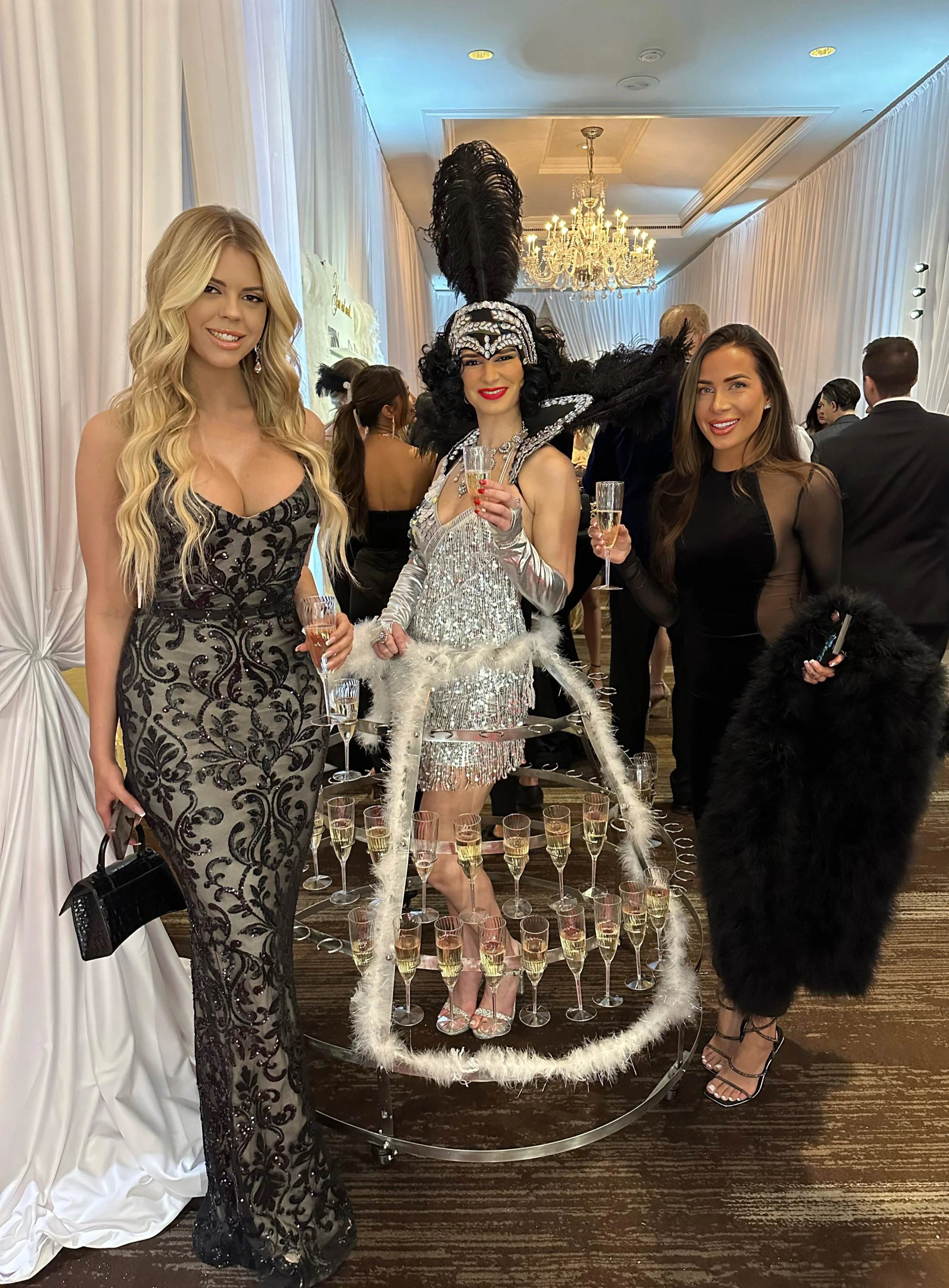 Three women at a glamorous event; one is dressed in a sparkling silver outfit with a feathered headpiece, standing inside a champagne glass display. The other two women wear elegant black dresses, holding champagne flutes. A chandelier hangs overhead