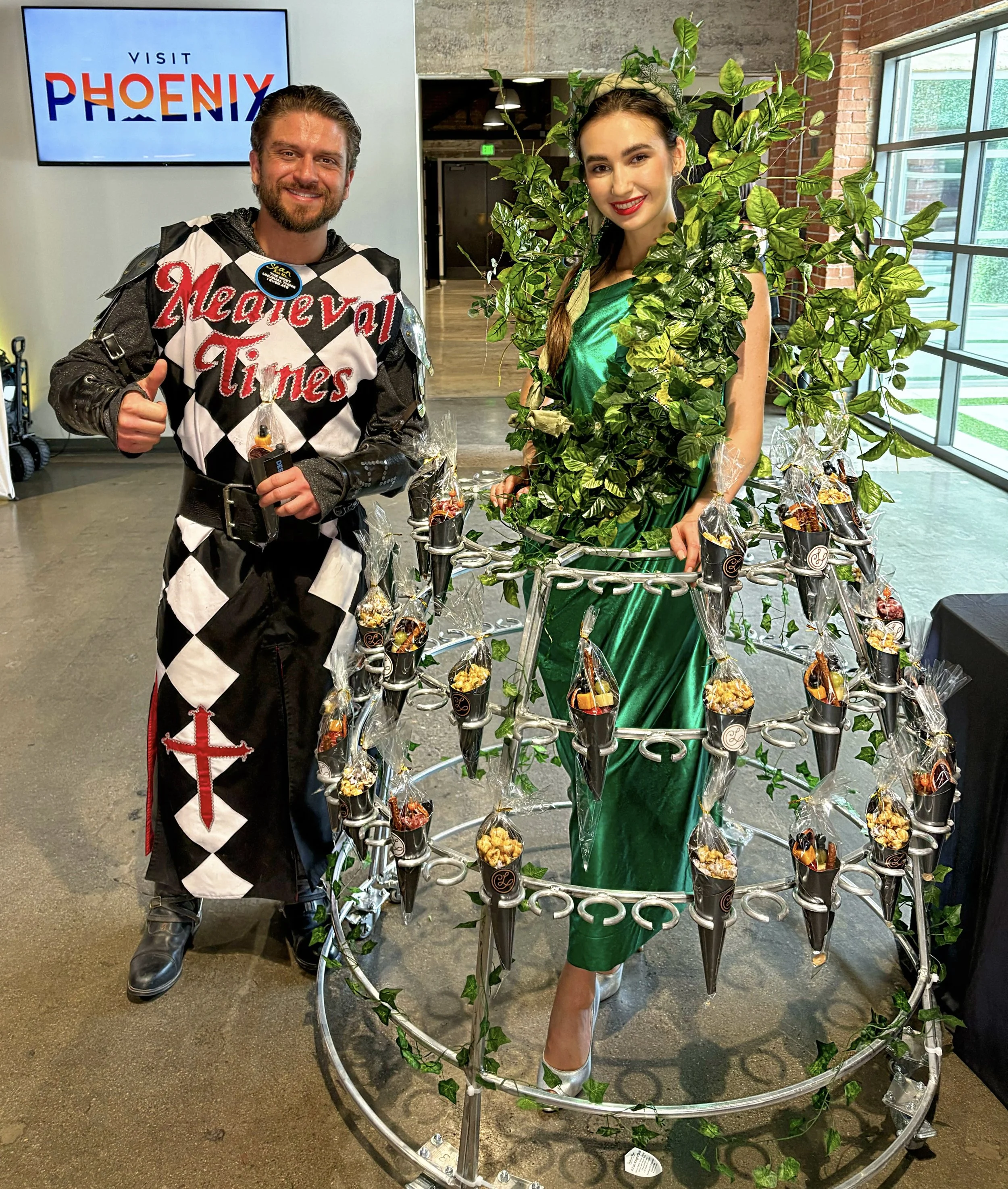 Two people posing indoors; a man in a Medieval Times costume and a woman dressed as a green plant with snacks attached to her outfit. A sign in the background reads 'Visit Phoenix.' The woman stands within a circular frame holding packaged snacks.