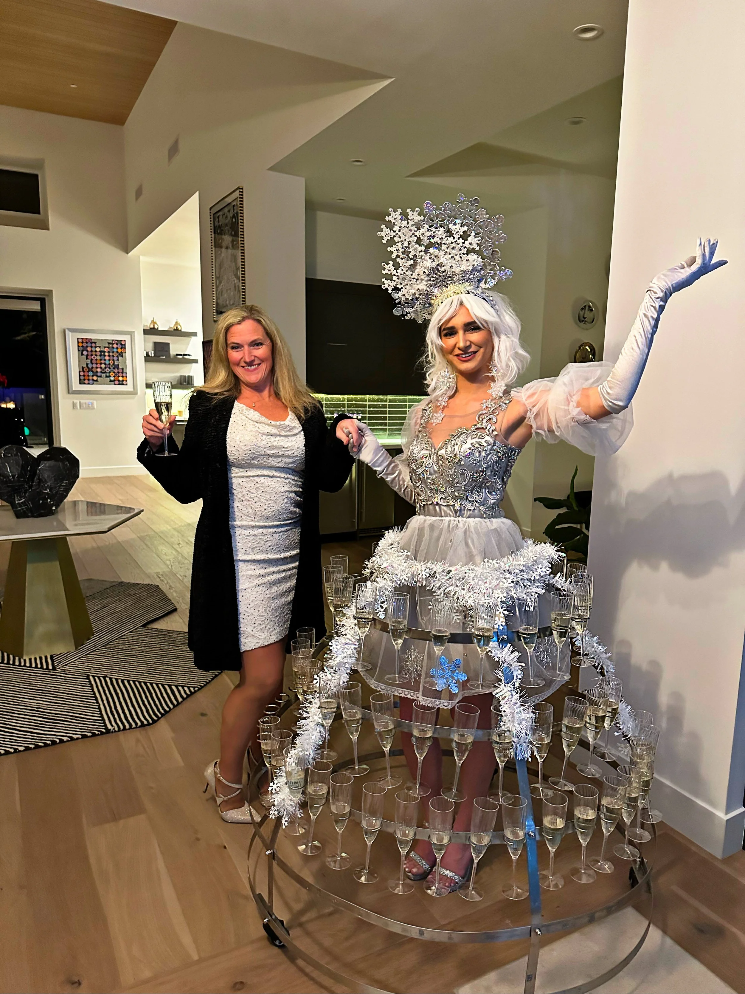 Woman in a white dress holding a drink next to another woman dressed in a silver and white costume resembling a champagne dress with glasses.