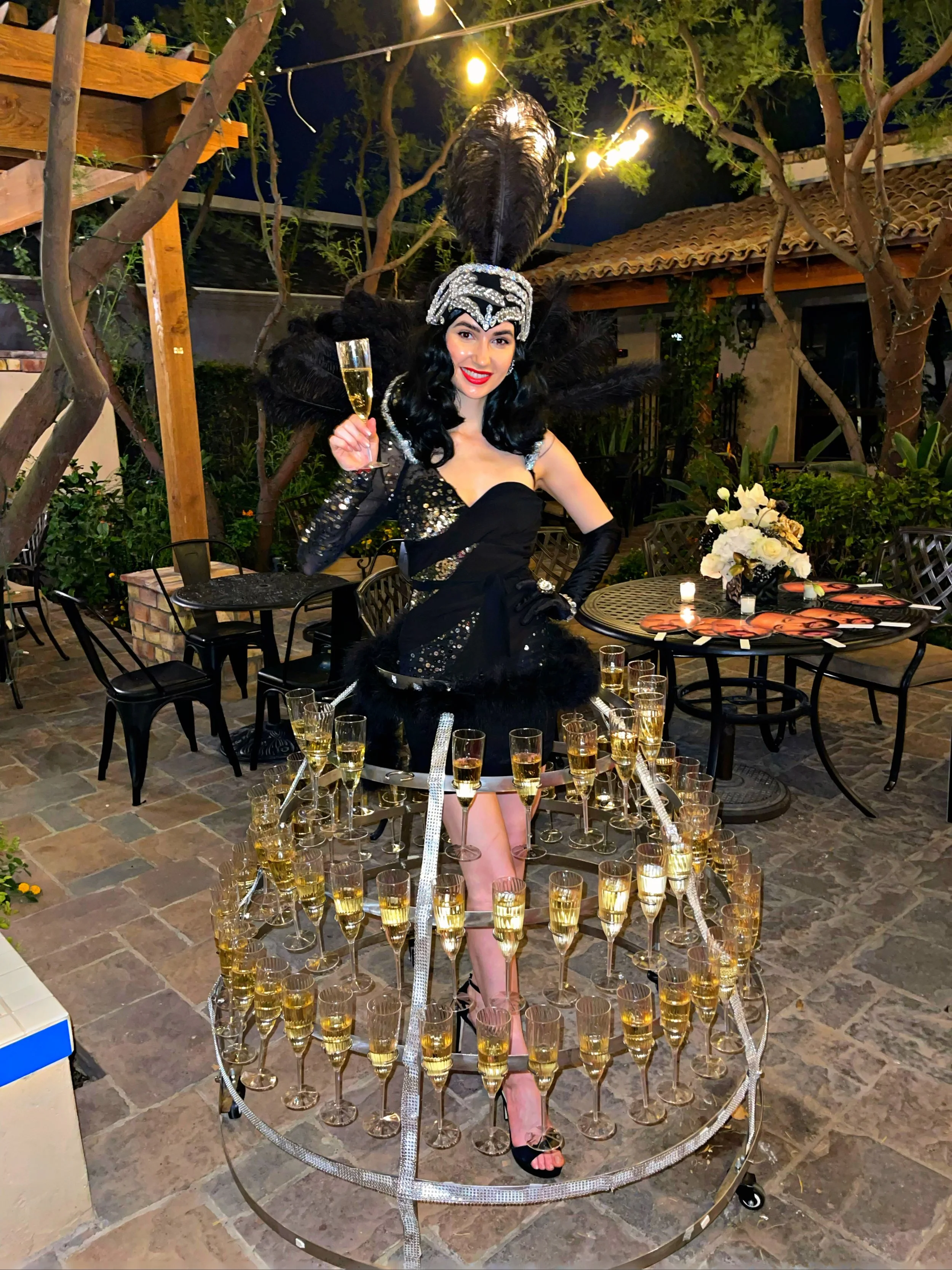 A woman dressed in a glamorous black outfit with a sequined headdress and feathered embellishments, holding a glass of champagne. She stands within a circular metal frame filled with multiple champagne glasses, set in an outdoor patio setting with tr