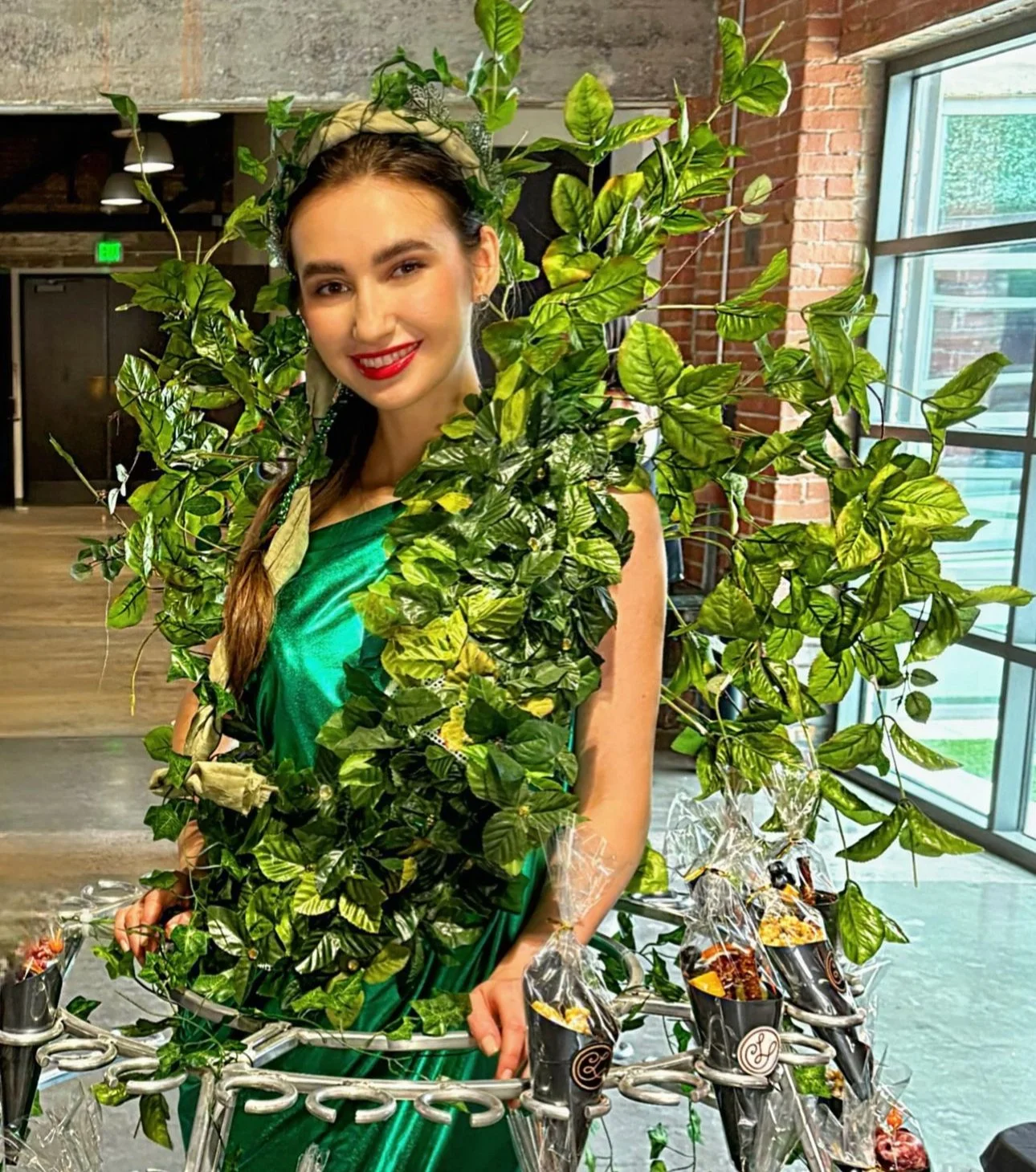 Person dressed in green with leaves, posing indoors with a decorative cart holding small wrapped items.