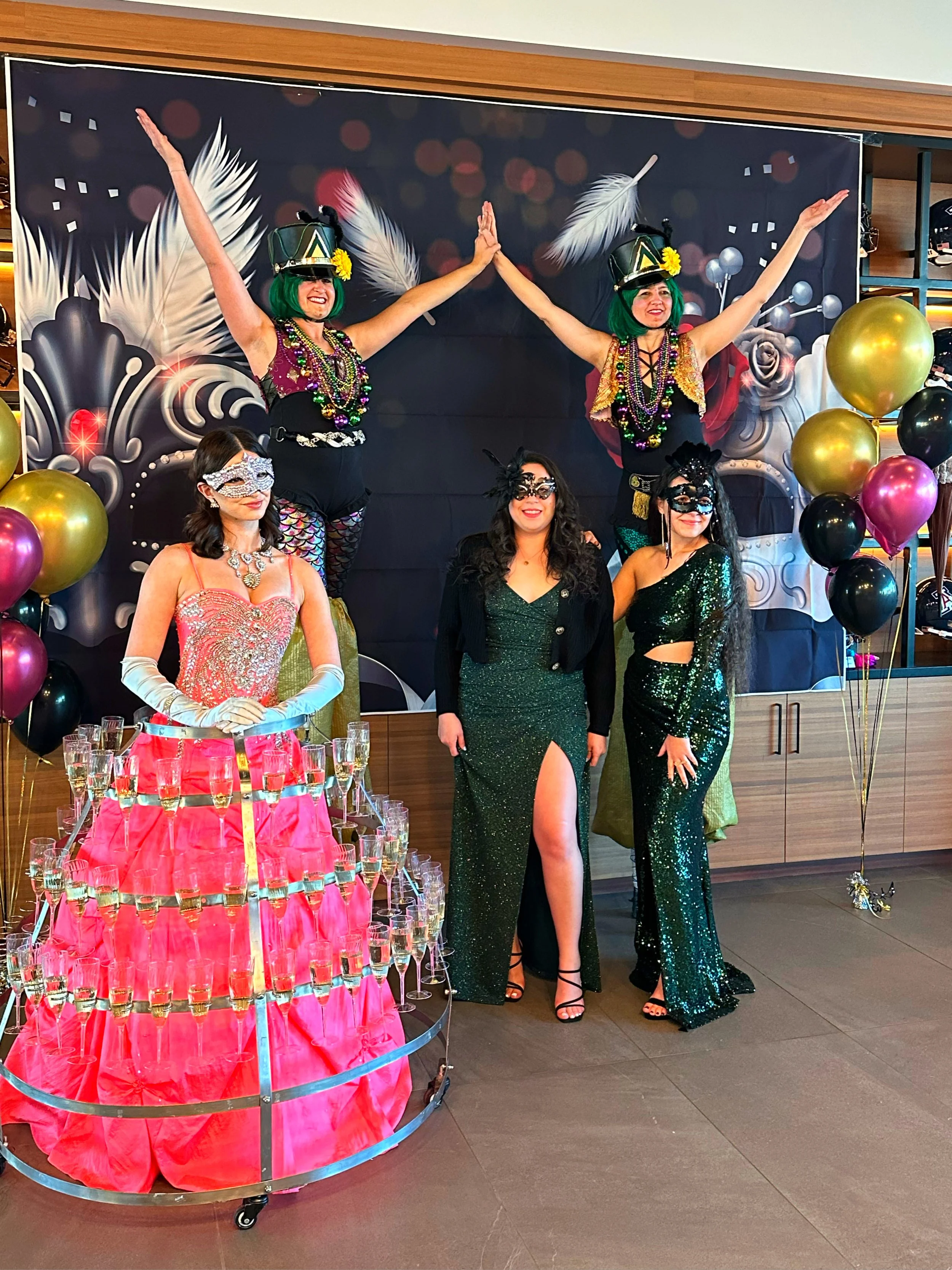 Group of women in costumes and masks at a festive event, with champagne glasses displayed on a skirt and balloons in the background.