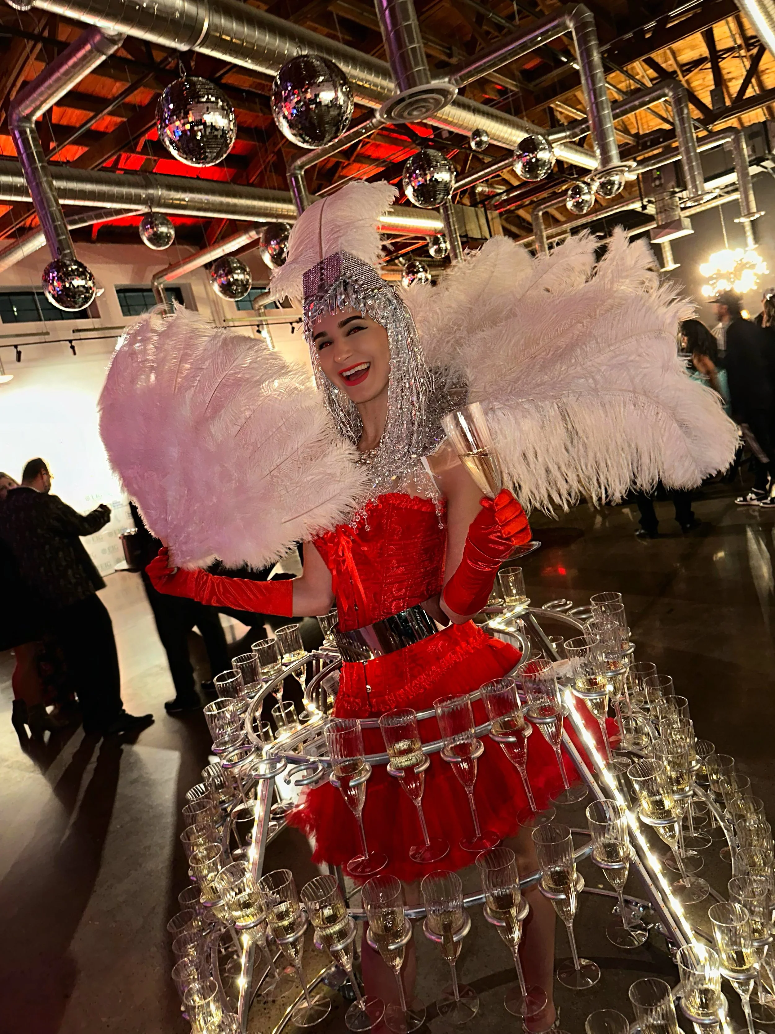 A performer wearing a silver headdress and red costume with white feathers, surrounded by a champagne glass holder at a party with disco balls.