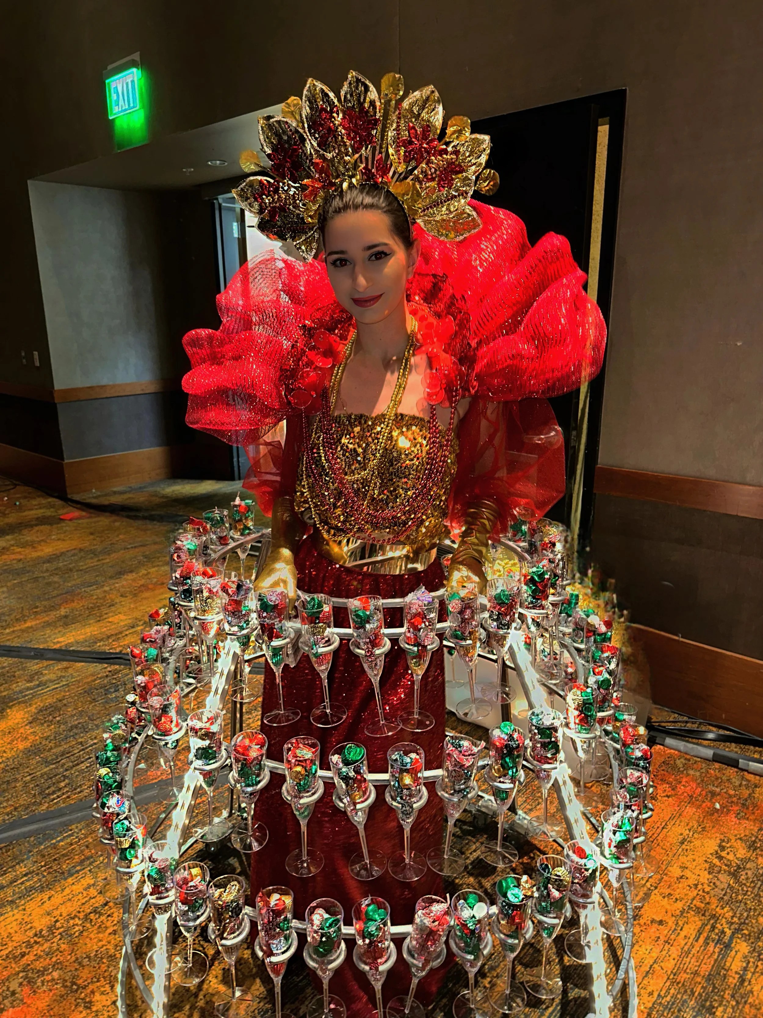 Person wearing an elaborate red and gold costume with a large headdress and wing-like shoulder pieces, surrounded by multiple tiers of filled glasses on a circular frame.