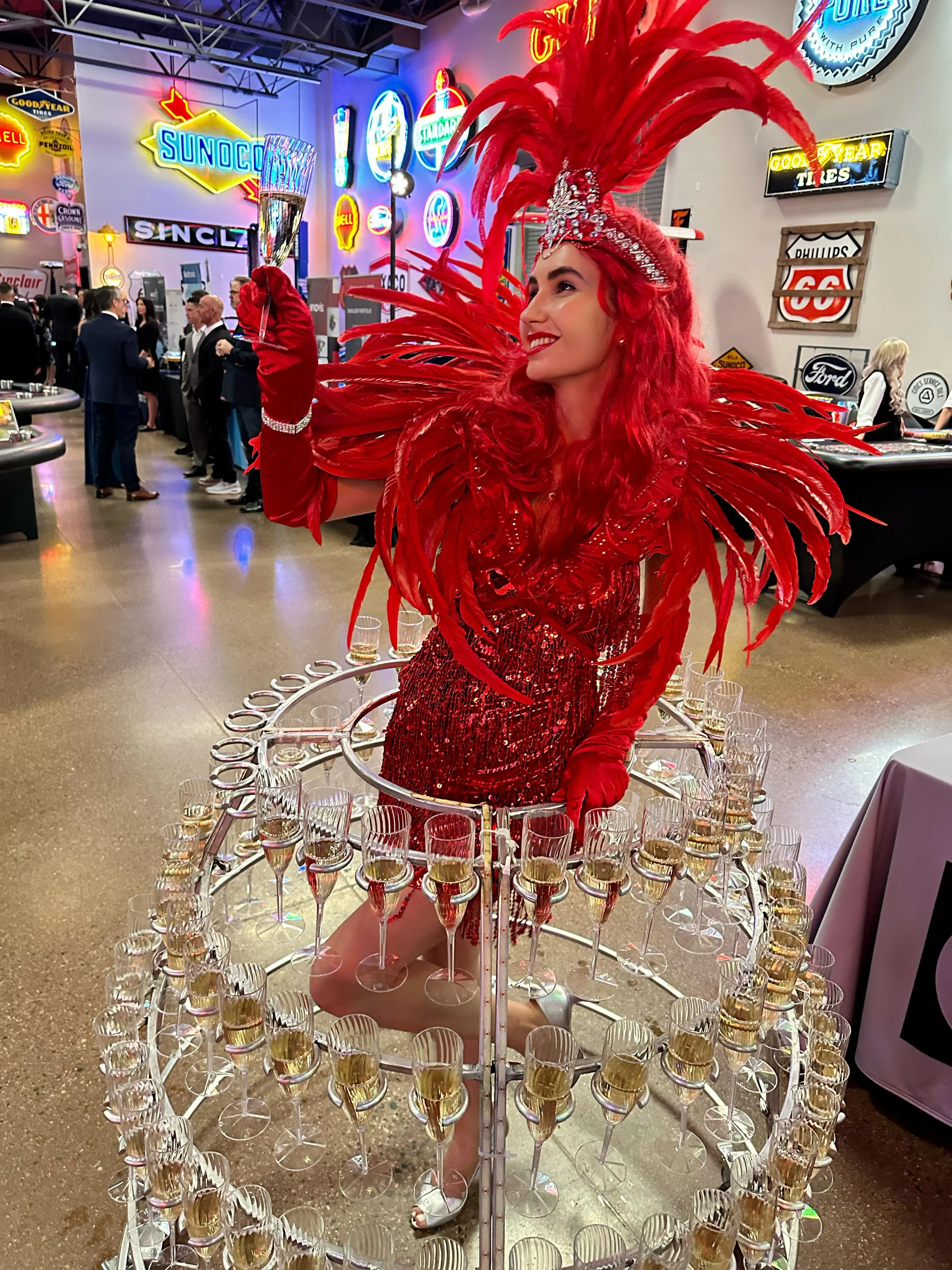 Woman dressed in red showgirl costume surrounded by champagne glasses in a neon-lit room.
