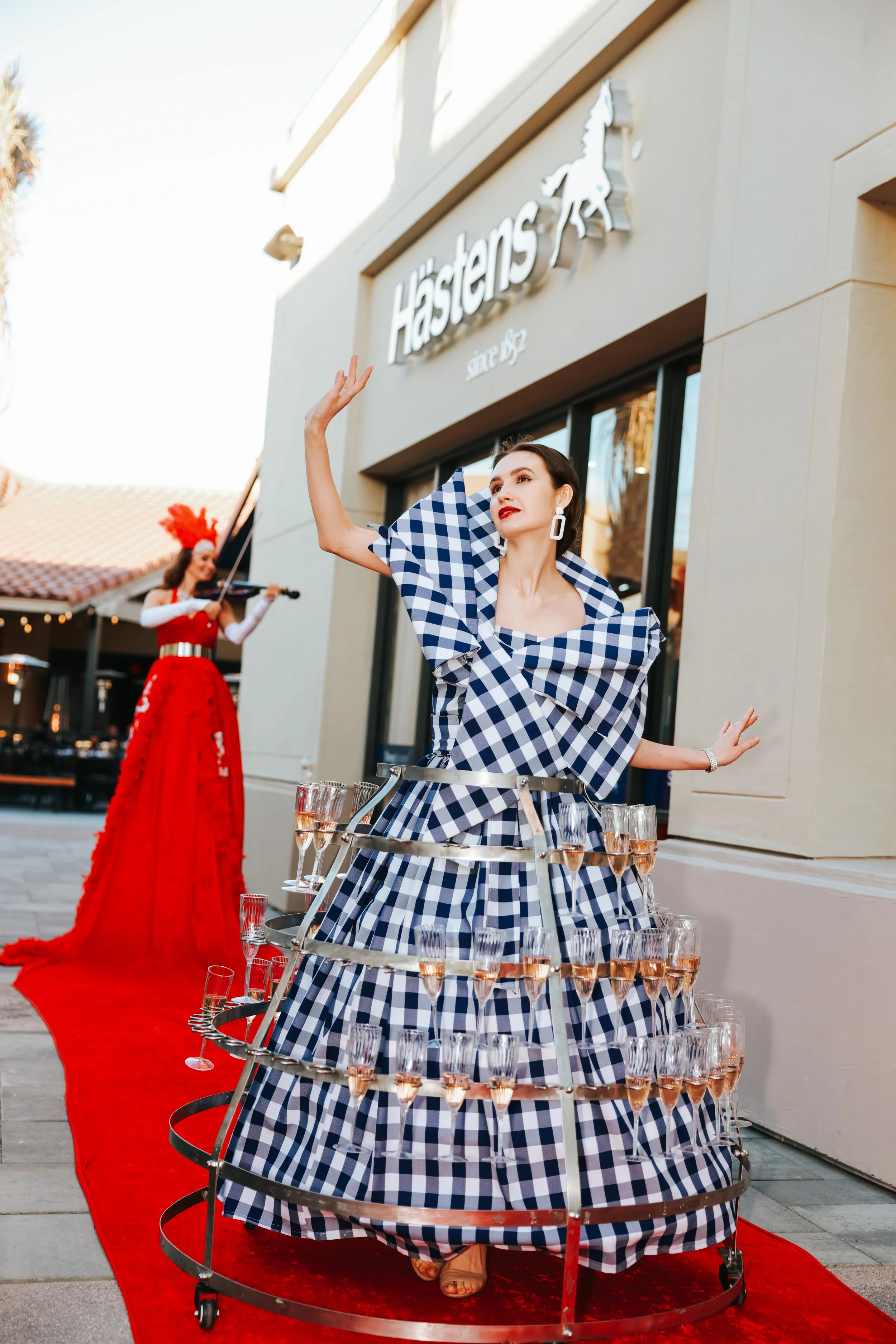 Woman in a blue and white checkered dress with champagne glasses, standing on a red carpet outside Hästens store, with a violinist in a red dress in the background.