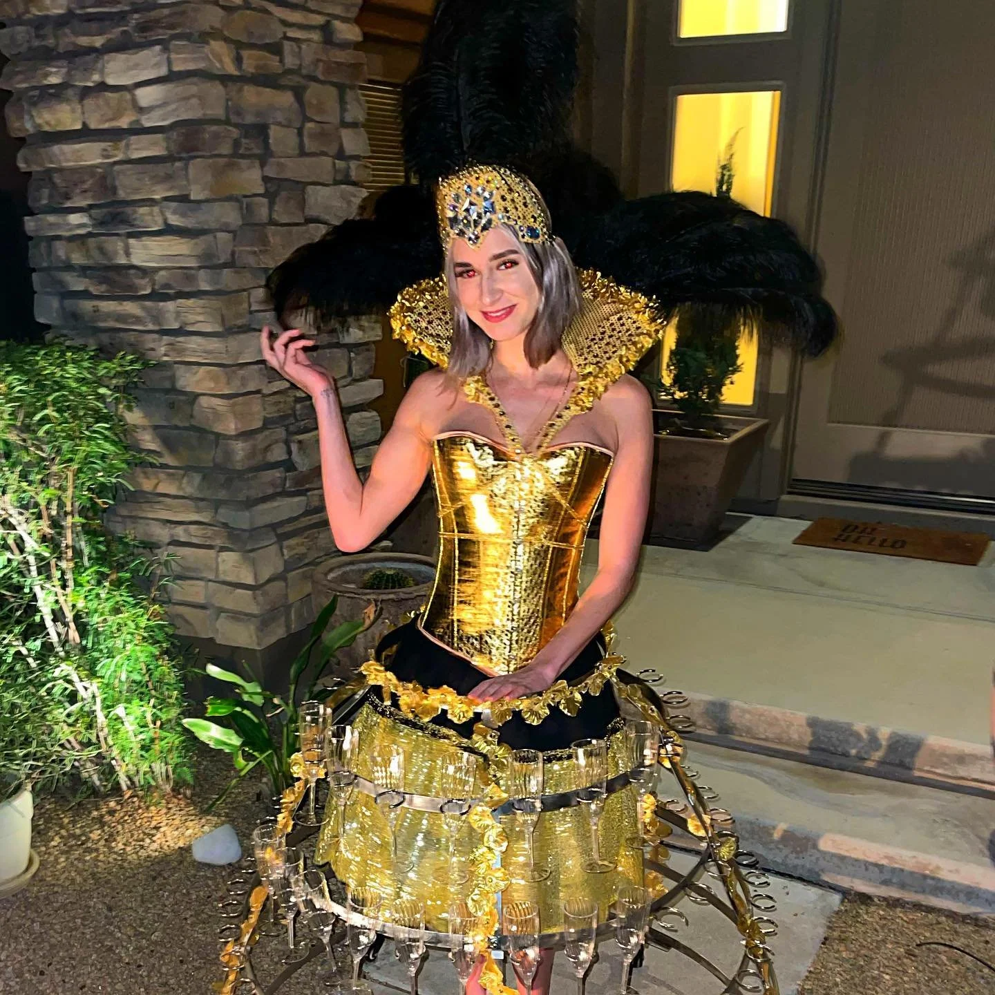 A person wearing a gold metallic costume with a corset and cage-like skirt adorned with glasses and black plumes, posing outdoors in front of a stone wall.