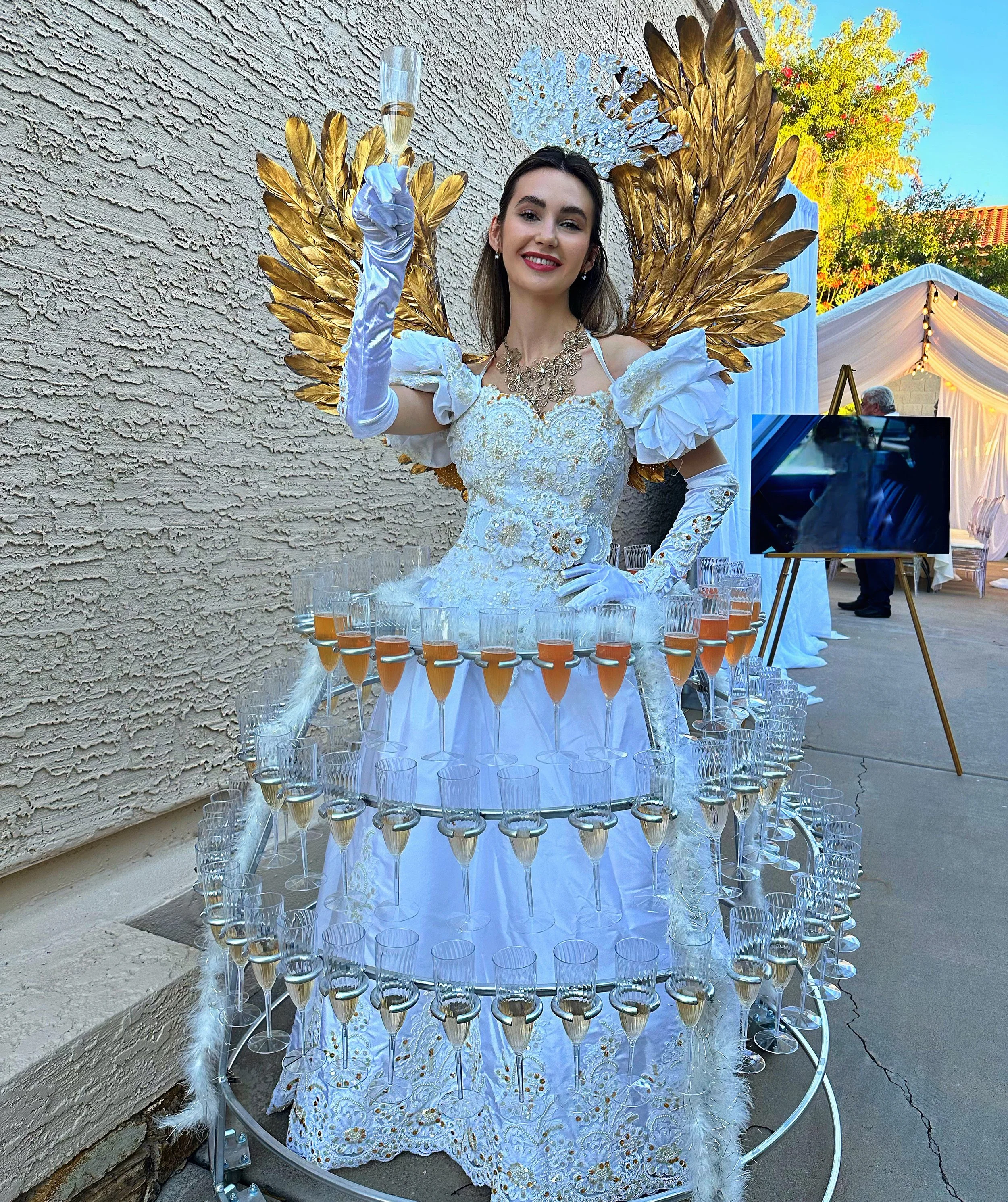 A woman wearing a white and gold decorative dress with golden wings and holding a glass of champagne. The dress includes multiple tiers with glasses of champagne.