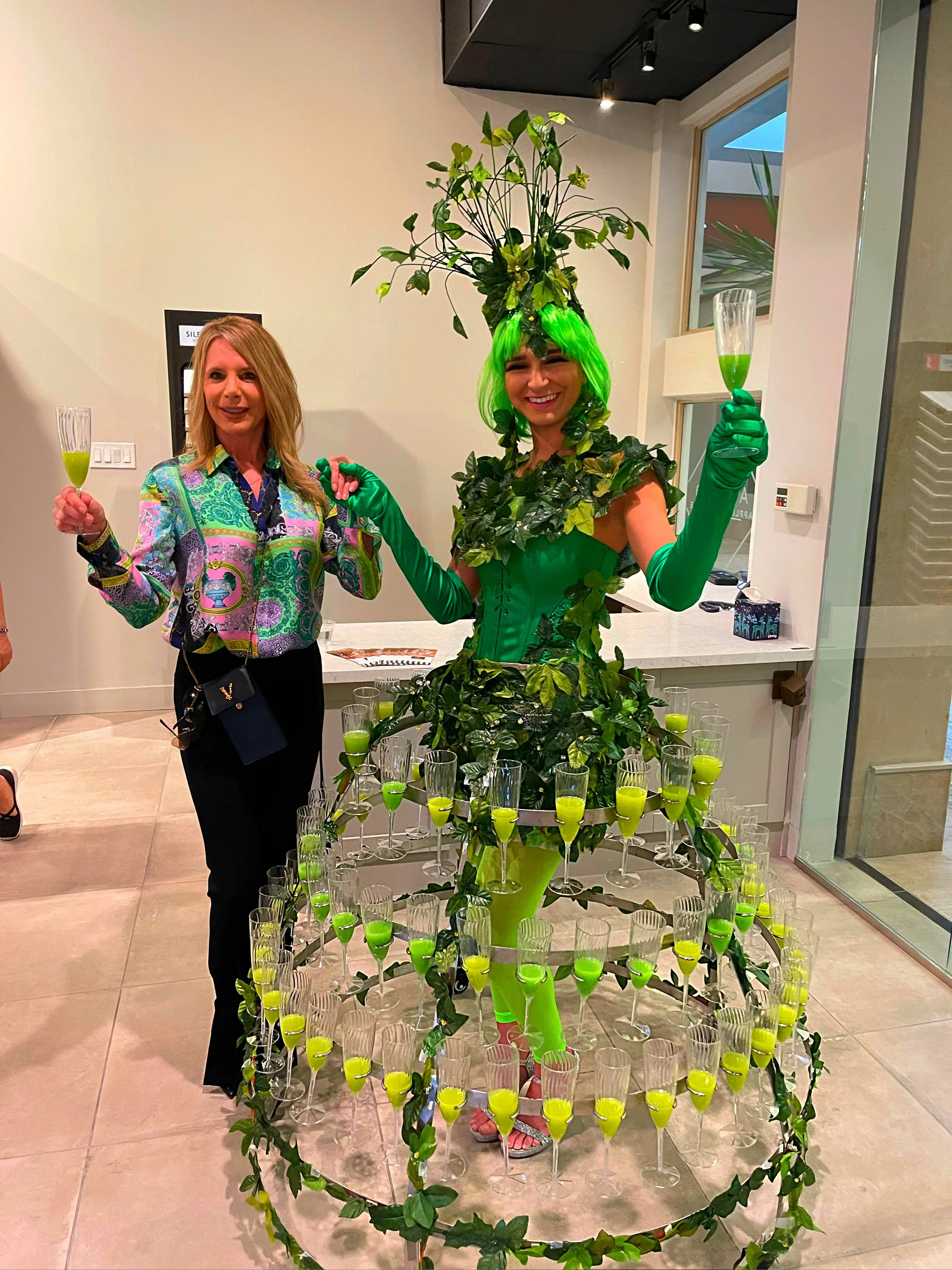 Woman in colorful dress holding a glass next to a performer in a green costume holding a similar glass, wearing a wig and foliage-themed hat, surrounded by a skirt-like tray holding multiple green drinks in glasses.