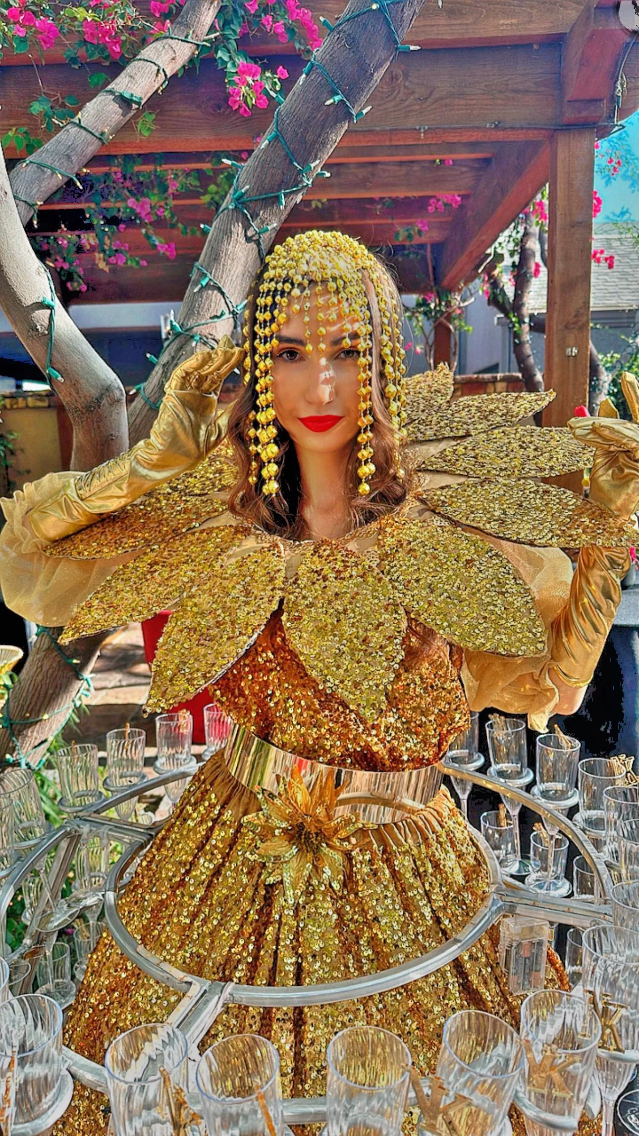 A person wearing a golden, sequined dress with leaf-like decorations, standing amidst a display of empty glasses. The person has a headpiece made of yellow beads and wears golden gloves. The backdrop includes wooden beams and green plants with pink f