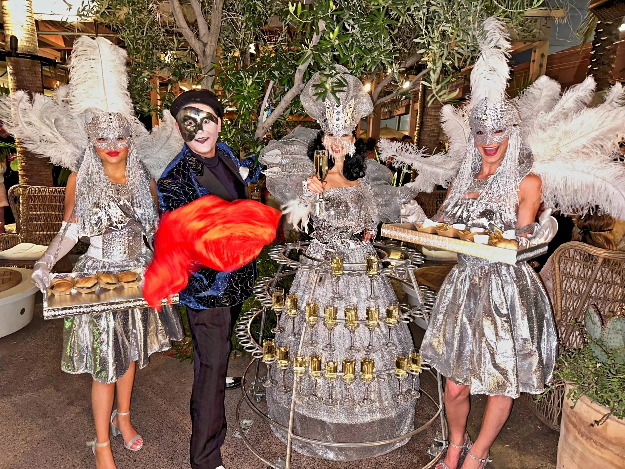People in elaborate masquerade costumes with silver and metallic attire. They have feathered headdresses and masks. The central figure wears a dress with champagne glasses attached. Others hold trays with hors d'oeuvres.